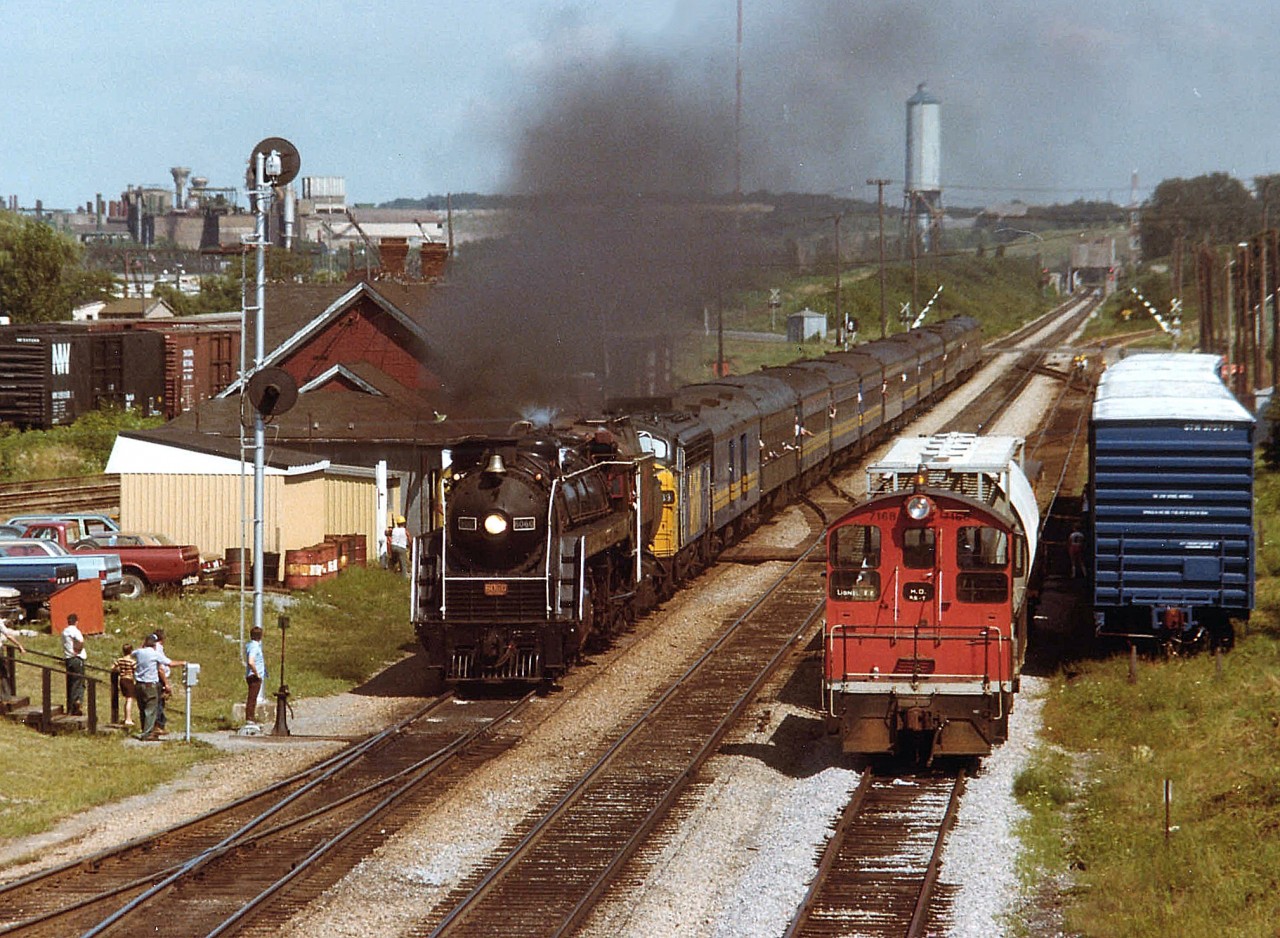 The days of CN's "Bullet-Nose Betty" 6060 getting rather long-in-the-tooth as she is now being assisted by VIA 6539 on the twice weekly tourist run from Toronto to Niagara Falls and return. In this view, one can see the Welland Canal bridge 6 counterweight in the background, and crossing gates returning to their upright position at the Glendale Av crossing, as the Toronto-bound afternoon train roars past the old Merritton station (destroyed by fire Oct 1994). Some of the local population watch in admiration. That is the old GM plant in background on the left; foreground freight cars are in now-taken-up Merritton yard, while local switcher CN 7168 waits it all out.
