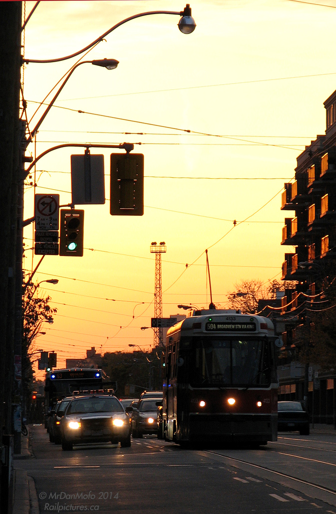 After taking on passengers, TTC CLRV 4133 pauses at the lights of King and Strachan before continuing on a 504 King run for the downtown core, in the gentrified and hip King West neighbourhood.  Once lined with garment factories and heavy manufacturing industry like the Toronto Carpet Factory and Massey Harris's equipment manufacturing plants (later Massey Ferguson), the departure of industry in the 80's and 90's coupled with the desire to live downtown saw this area along King Street converted from heavy industrial to commercial and residential use. Old factories were torn down for new condos and townhouses, with some older historic buildings repurposed into offices, lofts and residential units. The connecting route 63 bus behind 4133 will turn down Strachan for the trendy Liberty Village, to service streets that people once went to work along but now come home to. All the new residents in this area consequently have put a strain on transit, and the King streetcars are often packed to the doors during rush hour.  Another casualty of this gentrification can be seen looming in the background like a beacon in the sunset: one of the lighting towers for CPR's now-removed Parkdale Yard, sitting abandoned with nothing more to light. Time turned Parkdale Yard into townhouses, office buildings and retail outlets, as when all the rail-shipping industry moved out, so too did the need for a rail yard to serve them.