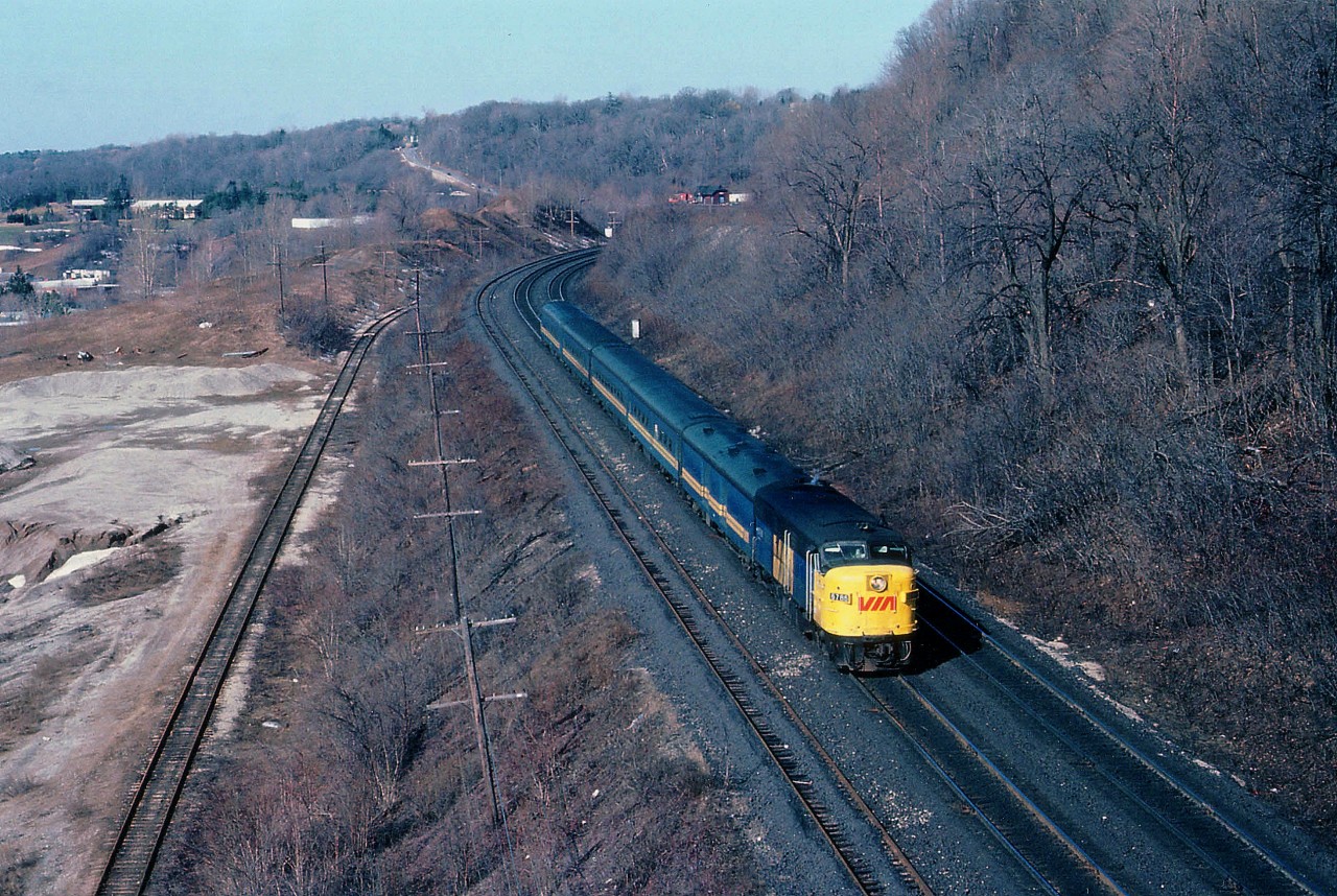 Beautiful March morning as #70 eastbound scoots down the grade toward Bayview Jct and destination Toronto. View is from the remains of the old Canada Crushed Stone Company conveyor overhang that until the '90s straddled the mainline. The two tracks on the left are gone now, the one by the double track CN main stored loads/empties for CCSL and the track down below was access track for Steetley, Inc., an operation just out of view on the left, at which dolomite was loaded for shipping via TH&B (which also had a connection thru Dundas to Aberdeen) as well as via the CN. A small industrial switcher shunted cars for this outfit. All is gone now, the access track last to go, taken up in August of 1985. A new subdivision of above average homes occupies this land today. Upper center in background of photo one can just make out Dundas station, surrounded by MoW equipment.