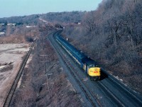 Beautiful March morning as #70 eastbound scoots down the grade toward Bayview Jct and destination Toronto. View is from the remains of the old Canada Crushed Stone Company conveyor overhang that until the '90s straddled the mainline. The two tracks on the left are gone now, the one by the double track CN main stored loads/empties for CCSL and the track down below was access track for Steetley, Inc., an operation just out of view on the left, at which dolomite was loaded for shipping via TH&B (which also had a connection thru Dundas to Aberdeen) as well as via the CN. A small industrial switcher shunted cars for this outfit. All is gone now, the access track last to go, taken up in August of 1985. A new subdivision of above average homes occupies this land today. Upper center in background of photo one can just make out Dundas station, surrounded by MoW equipment.