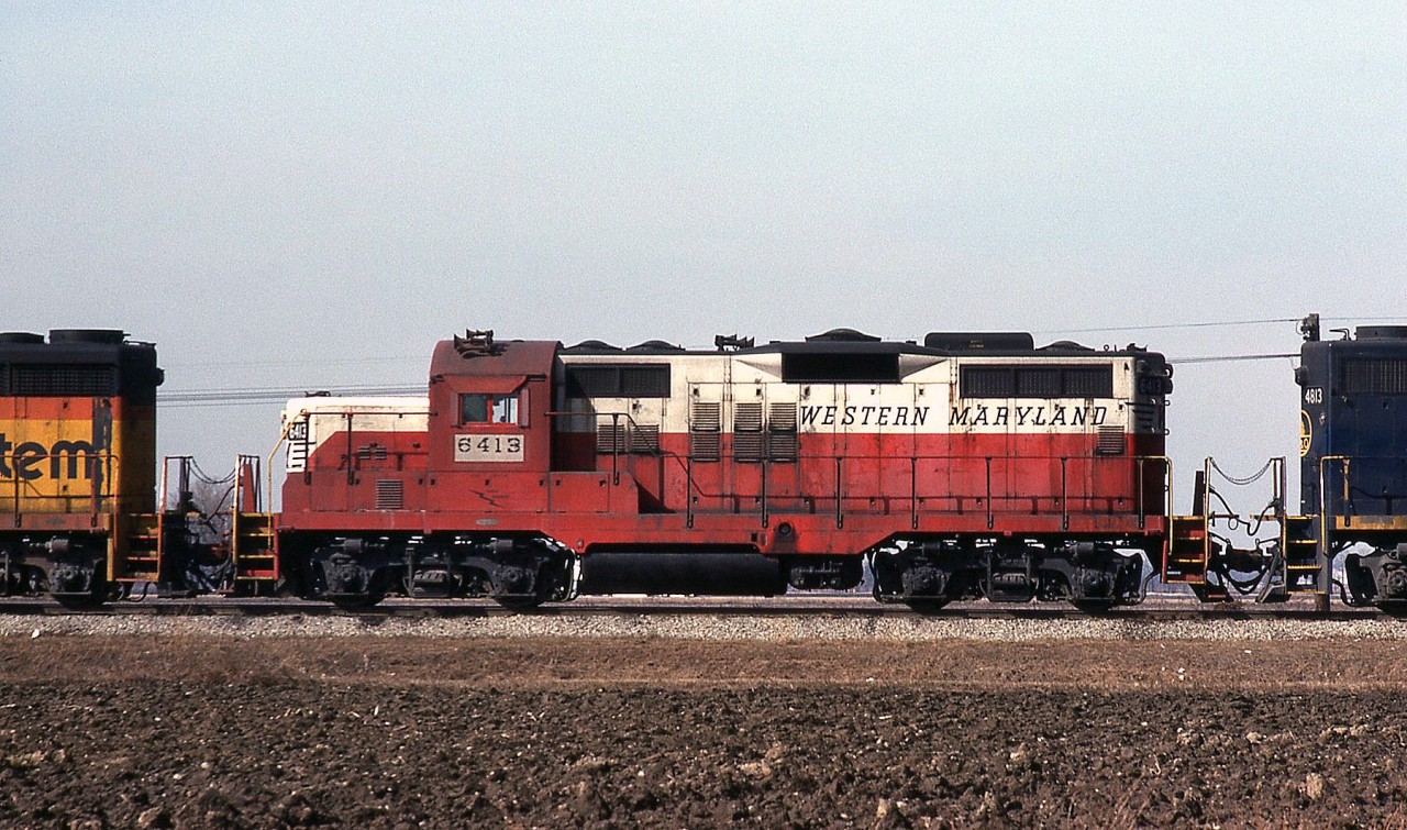 Western Maryland GP9 6413 is shown in-consist trailing a Chesapeake & Ohio train near Oldcastle ON on C&O's Subdivision 1. WM gave their early Geeps a taller chop-nose job than most, giving them a bit of an odd appearance that some will either love or hate. The unit in front is a GP30 or 35 in the Chessie System livery, the trailing unit 4813 a GP38 in B&O's blue dip. WM was absorbed into the B&O 4 years ago in 1983, and while some units were repainted into the Chessie System livery with WM reporting marks, 6413 still retains the WM's old "Circus colours" livery. 

At this point in 1987 operations were still as the Chesapeake & Ohio Railway, albeit under the Chessie System banner (since 1973). Subsidiary B&O would be folded into the C&O less than two months later on April 30th. Parent CSX Corporation would officially absorb the C&O Railroad into their new railroad division, CSX Transportation, a few months after on August 31st.

Note: Geotagged location not exact.