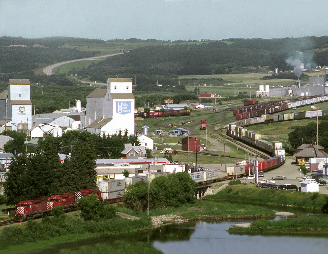 Eastbound Edmonton to Winnipeg Containers train 464 running on the North Main starts up the 2 percent grade out of the Little Saskatchewan valley after a crew change at the station visible on the right. The line west out of the valley also has a section of 2 pc grades