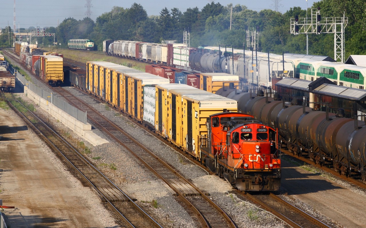 CN GP9RM 7075 and GMD-1 1412 wait to clear track 32 to the lower yard as 399 begins to back 22 autoracks for Ford onto track 30 (off screen to the right). The rest of 399 is sitting on main track one in the background beside the Hamilton bound GO train at the station on main track two. Way in the background the westbound L-10-L GO train leave Aldershot on main track three. Rush hour is a bus time for the dispatcher, which translates into entertainment for railfans!