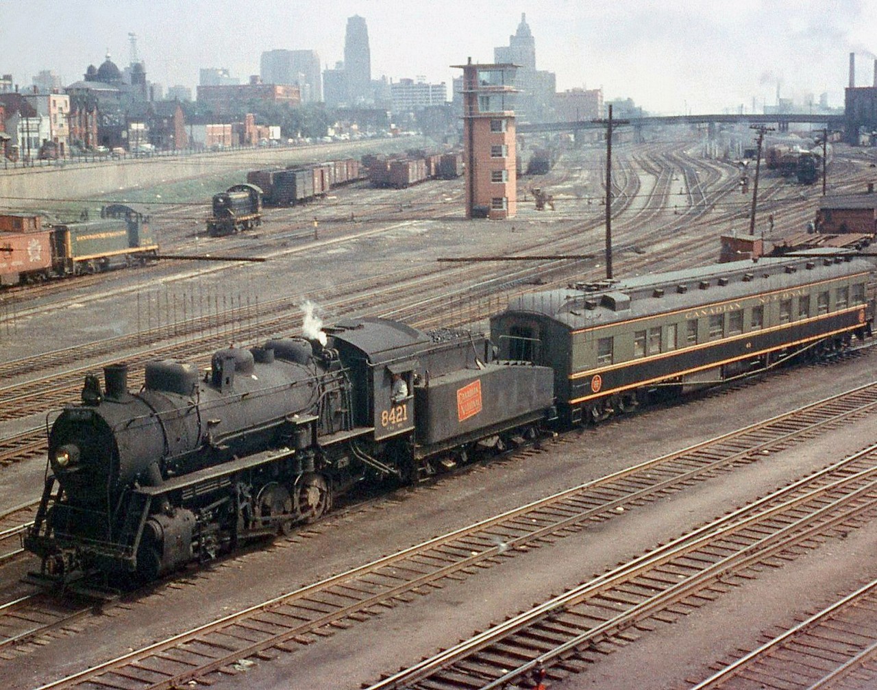 Steam era action at Bathurst Street: Canadian National 0-8-0 8421 switches a heavyweight business car around the Spadina Coachyard (off to the right) at Bathurst Street bridge. New GMD SW1200RS and ALCO/MLW S-series diesel switchers work the freight-only Bathurst Street yards to the left. The CN Spadina Roundhouses' coaling tower is visible to the upper right, where dozens of steam and diesel power were maintained for service out of downtown Toronto. Also of note is the relatively new looking brick yard tower (identical ones were at Mimico and Don Yard, all demolished today). The Toronto skyline 50+ years ago looks much different than it does today, with the tallest building being the Canadian Commerce Bank Building (which was also the tallest building in the British Commonwealth). It held this distinction from 1931 to 1962, and was 69 feet taller than the Canadian Pacific's Royal York Hotel, also visible here above the yard tower.  CNR (or GTW) 8421 shown here was one of six P5j 0-8-0 switch engines built by Brooks, purchased from original owner Buffalo Creek Railroad in 1947 and assigned Grand Trunk 8400-series numbers. "GTW" 8421 (however it's shown here sporting CN lettering on the tender's wafer logo) is former BCK 27, and operated in GTW/CN service with its former owner's number and lettering after purchase until becoming 8421. Most photos show these steamers around downtown Toronto, switching the freight and passenger yards in the area. All were retired by the end of the steam era in Canada, and scrapped.