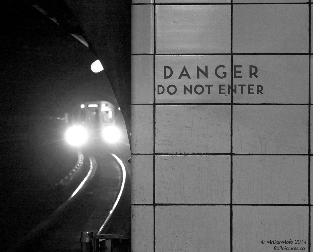 As the lead H6 car rounds the bend with lights glaring, invoking images of the original "The Taking of Pelham 1-2-3" movie posters (the famed 1974 classic with Walter Matthau and Robert Shaw), sans gun-toting hijackers of course, a TTC subway train arrives westbound at Yonge Subway Station on the Bloor-Danforth line. The warning etched in the dirty wall tiles underscores the dark dangers lurking in the tunnels, from a high-voltage electrified third rail to close quarters and minimal side clearances for any trespassers.