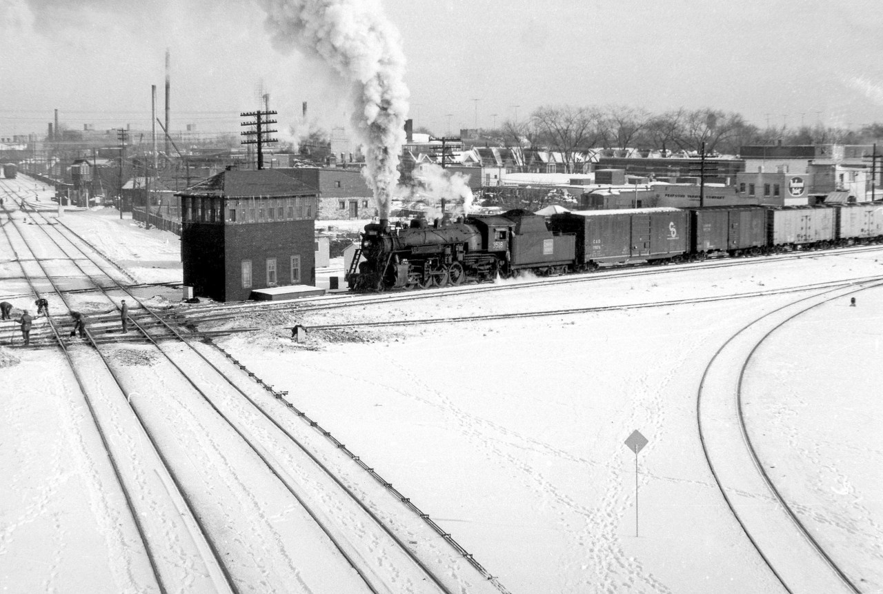 Steam-era West Toronto Diamond in the Winter: Canadian National Mikado 3518 heads north on the Brampton Sub (today the GO Weston Sub), passing the former West Toronto interlocking tower and about to cross the diamonds with the CP's North Toronto Sub. A group of employees tend to the ten(!) diamonds that make up this interlocking. In the background on the left one can spot the CP grade crossing tower for Osler Avenue. On the right, the tracks of the CP Galt Sub curve under the now-demolished Old Weston Road bridge where this photo was taken from.  The large human-manned interlocking tower here locally controlled the passage of CN & CP trains over each others' tracks, and was demolished around when the interlocking plant was upgraded to be remotely control by the CP Toronto Terminal RTC around 1965 (photo of the area soon after here). In May 2014, the final train crossed the diamonds here - after years of construction work, GO Transit/Metrolinx converted the diamonds to a fly-under to reduce delays for increasing passenger service on this corridor.  The closest pair of diamonds are for the CP Galt-MacTier connecting track, allowing trains like The Canadian heading to and from nearby West Toronto Station to get onto the MacTier Sub (shown here by James Adeney using the connecting track). It also crossed over both tracks of the Brampton Sub out of frame to the left via two more diamonds. The next two tracks are the afforementioned CN Brampton Sub mainline from downtown Toronto to Brampton, Guelph and beyond (later becoming the Weston Sub, today owned by GO Transit).  The last track crossing just behind the tower is CP's "old Bruce" service track to industries on the east side of the corridor, heading as far down as Laura Secord (Nestle today) at Dundas Street before connecting with CN near Parkdale. It was part of the original Toronto, Grey & Bruce line before CP absorbed it, with much of the line to the north becoming the CP's MacTier Sub. The Penfound Varnish Company on the right was also serviced off this track. This diamond crossing was eventually removed and replaced with a sharp curving lead starting by Osler Ave. The old Bruce service track was fully removed in the 90's after much of the manufacturing industry along this corridor located elsewhere or stopped shipping via local sidings. Today all the industry and customers that once shipped by rail in this area are gone, but mainline freights, passenger and commuter trains still pass through on a constant basis.