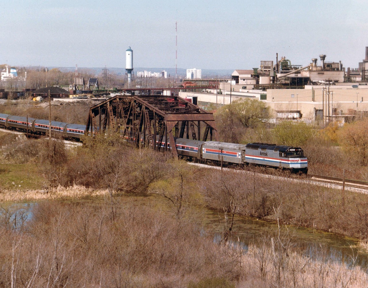 This is the First Run of the Maple Leaf thru Niagara over the CN Grimsby Sub. The train is shown eastbound crossing the trestle known on the timetable as "CN Iron Bridge", with the GM #2 plant dominating the background. On the left is the Welland Canal Lift Bridge #6, and the superstructure of a Lake Erie -bound ship can be seen. This photo was shot from up in now off-limits property of Walker Industries quarry, and is an impossible angle these days. Because this is a first run, the total consist was noted: AMTK Loco 344, baggage, 1370 and coaches 21126, 21148, 21071, 21054, 20224 and 21107.