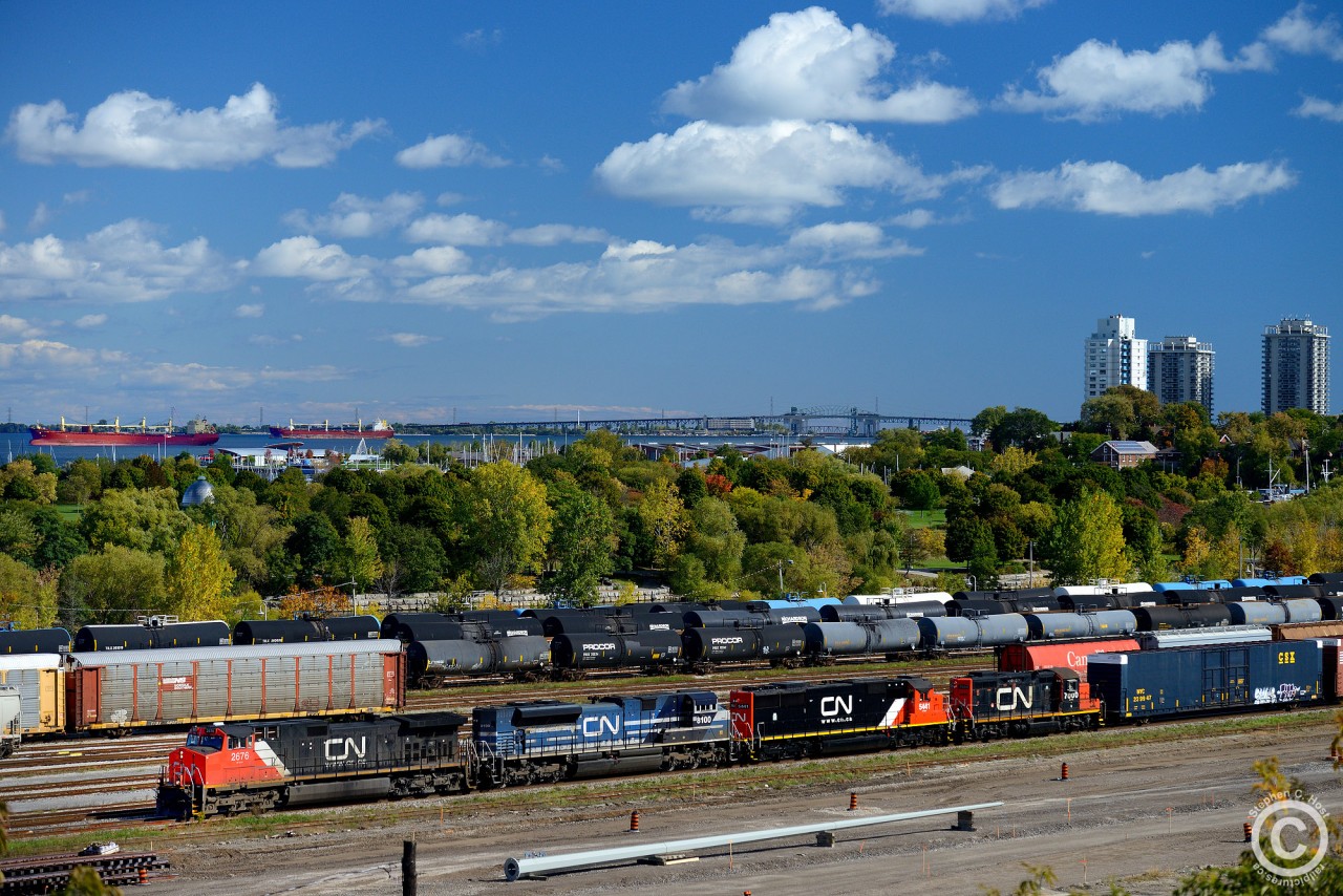 CN 422, with engines CN 2676, CN 8100, CN 5441, CN 7000 roll on the mainline of the Grimsby/Oakville connecting track with the rest of Hamilton yard in the background. Two vessels are seen in Hamilton Harbour waiting for a port berth, Heloise and Federal Elbe. Heloise docked at Richardson's terminal near the QEW, and Federal Elbe  at Federal Marine Terminals dock in Hamilton. Photo notes: used a polarizer filter.