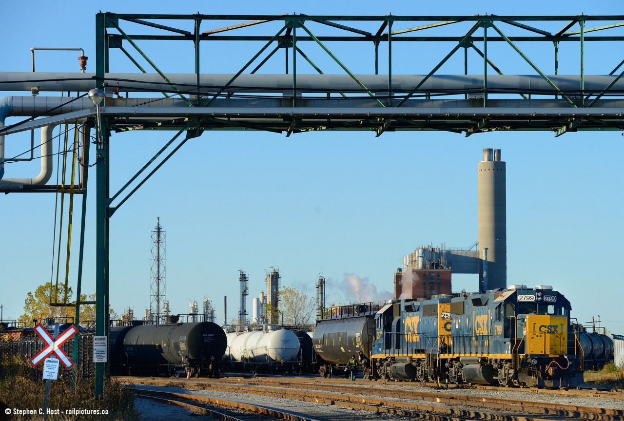 CSXT GP38-2 remote control mother 2799 is switching cars in the north end of the main Sarnia yard and is Framed in a pedestrian/pipeline overpass (long disused for pedestrians, but used for Imperial Oil pipelines). In the background is the idle Imperial Oil backup power plant.