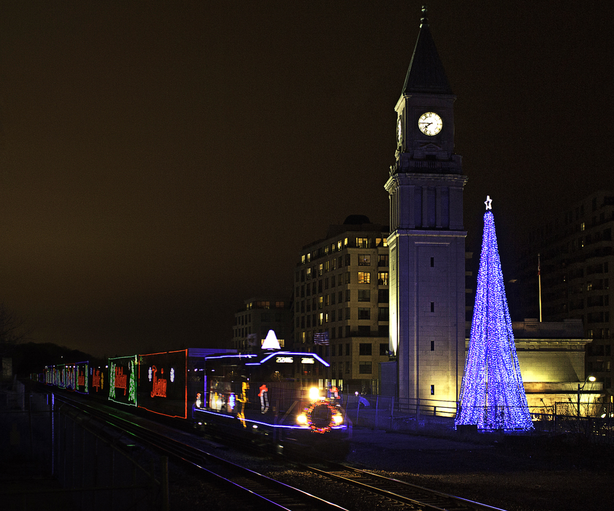 Lets just say, I was a bit high off the ground, and it was necessary to watch my step. An injured elbow was further harmed in the making of this photo. Here, the Canadian version of the Holiday train zips past the old North Toronto-Summerhill CPR station, with the now 9 year old Christmas tree adding to the scene. Hoping it would perform its slow rollby past Summerhill as it had done many years past, this year it zipped by at a good clip. Realizing a higher shutter speed would be detrimental to the lighting of the photo, I kept my shutter low sacrificing a slightly blurred leader. Having taken multiple low exposures beforehand, I thought adding a ghost effect would help compensate for the blur as well as add some Christmas magic. The Holiday train, introduced in 1999, has gone through a change of power three times. Starting out with SD9043MAC's and AC4400CW's, the AC4400CW took a primary role since until 2013. Being a loosely timed trip with a rather short train (for freight standards), newly rebuilt (ex GP9u's) GP20C-ECO's 2246 and 2249 have taken this year's role. Rolling by at 7:45PM, according to the clock tower, 01H would easily make it's next stop, Lambton yard by 8:15PM. As for the Christmas tree, it was first put up in 2005, probably for that year's Holiday train, which stopped at Summerhill that year. That was the one and only year it stopped at Summerhill, however the tree was still put up every year since. Slow rollby's, and happy horn blasts continued until recently. The importance of the location is not as recognized now, with this year's train passing at around 35mph, and the horn remained silent. Unless 02H will be doing this year's honours, but I think it's likely it'll rip through at track speed.