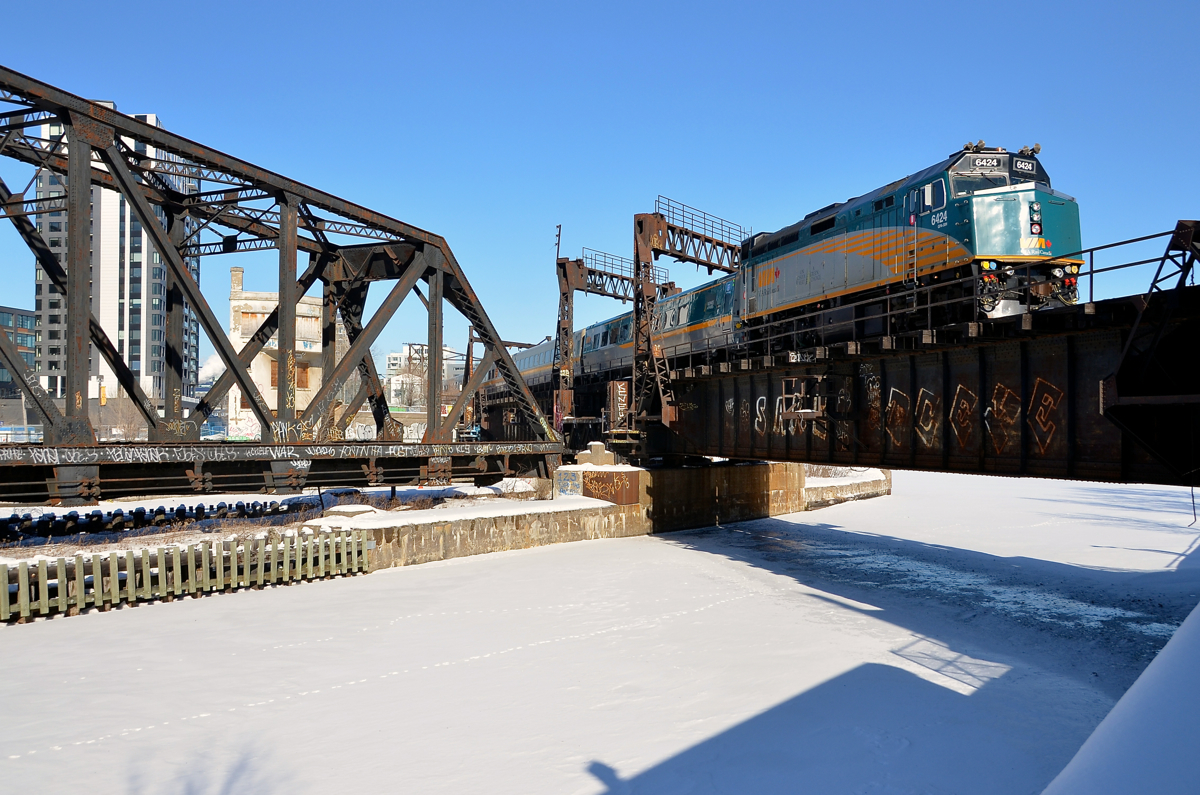 Backing up over the Lachine canal. VIA 6424 is backing up, pushing a deadhead movement towards Montreal's Central Station as it crosses the Lachine canal. Across the canal, on the other side of the out of use swing bridge, one can see the out of use Wellington tower.