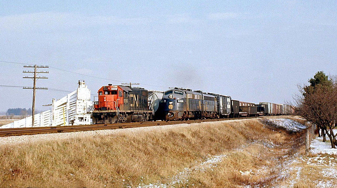 Canadian National GP38-2 5506 in in the siding by the co-op cattle pens with its train, as Norfolk and Western F7A's 3726 and 3666 pass by on the north main track at Thamesville on the CN Chatham Sub, likely after coming off the Paynes Sub to the east at Glencoe.