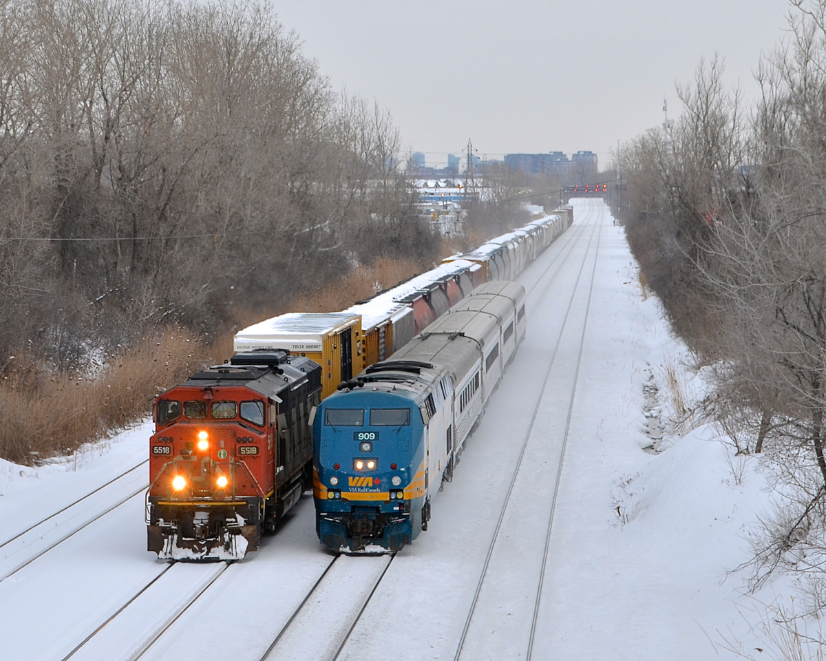 Both CN 527 (with CN 5518) and VIA 67 (with VIA 909) are moving as VIA 67 prepares to overtake CN 527, which is only doing a few miles per hour. Soon CN 527 will enter nearby Taschereau Yard.