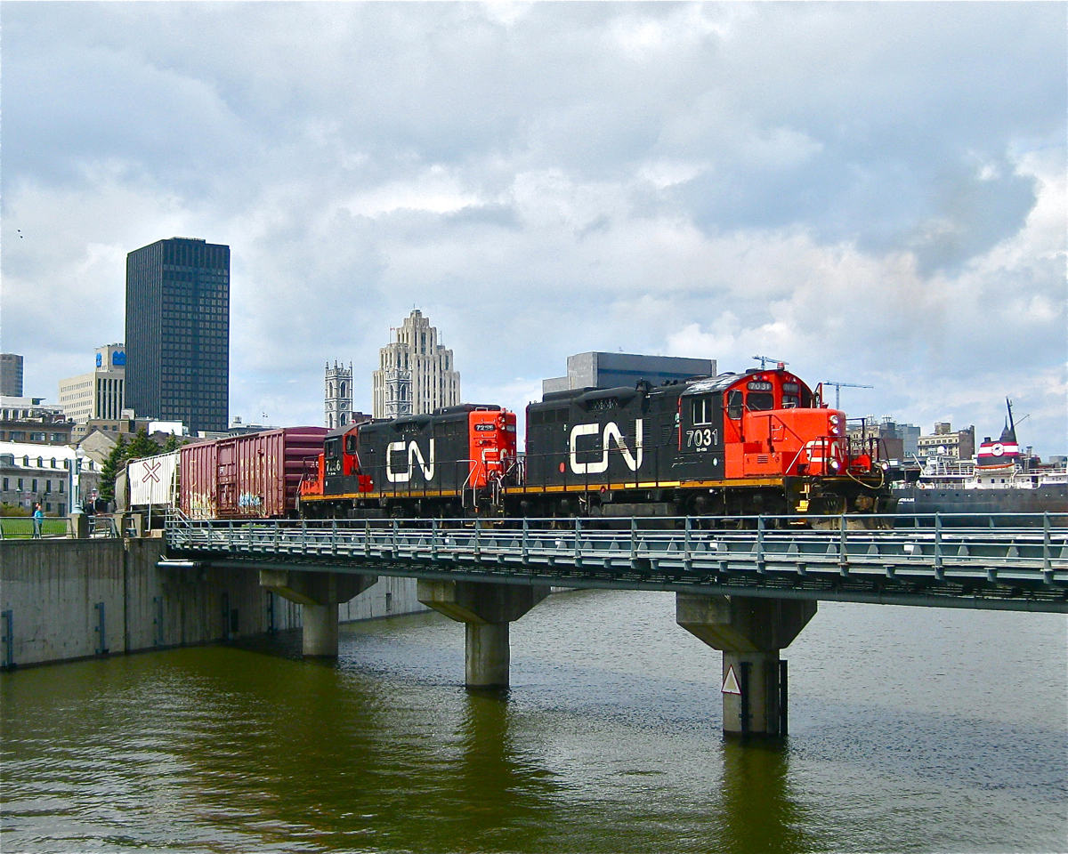 CN 7031 and CN 7256 shove a decent sized cut of grain cars east through the old port of Montreal. The train is crossing over the eastern end of the Lachine Canal and is about to curve to the east. In the distance at right is the ship "Algoma Montrealais".