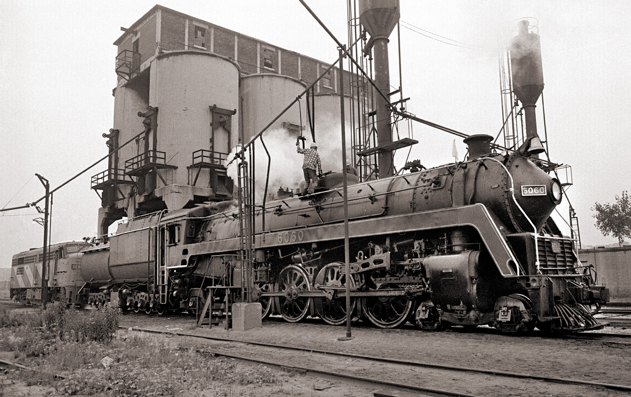 After one of the last CN steam excursions to Fort Erie, CN 6060 is serviced before returning to the roundhouse.  A shopworker climbs on top of the boiler to fill the sand dome.  The old coaling tower looms over the servicing tracks, although it hasn't been used for years.