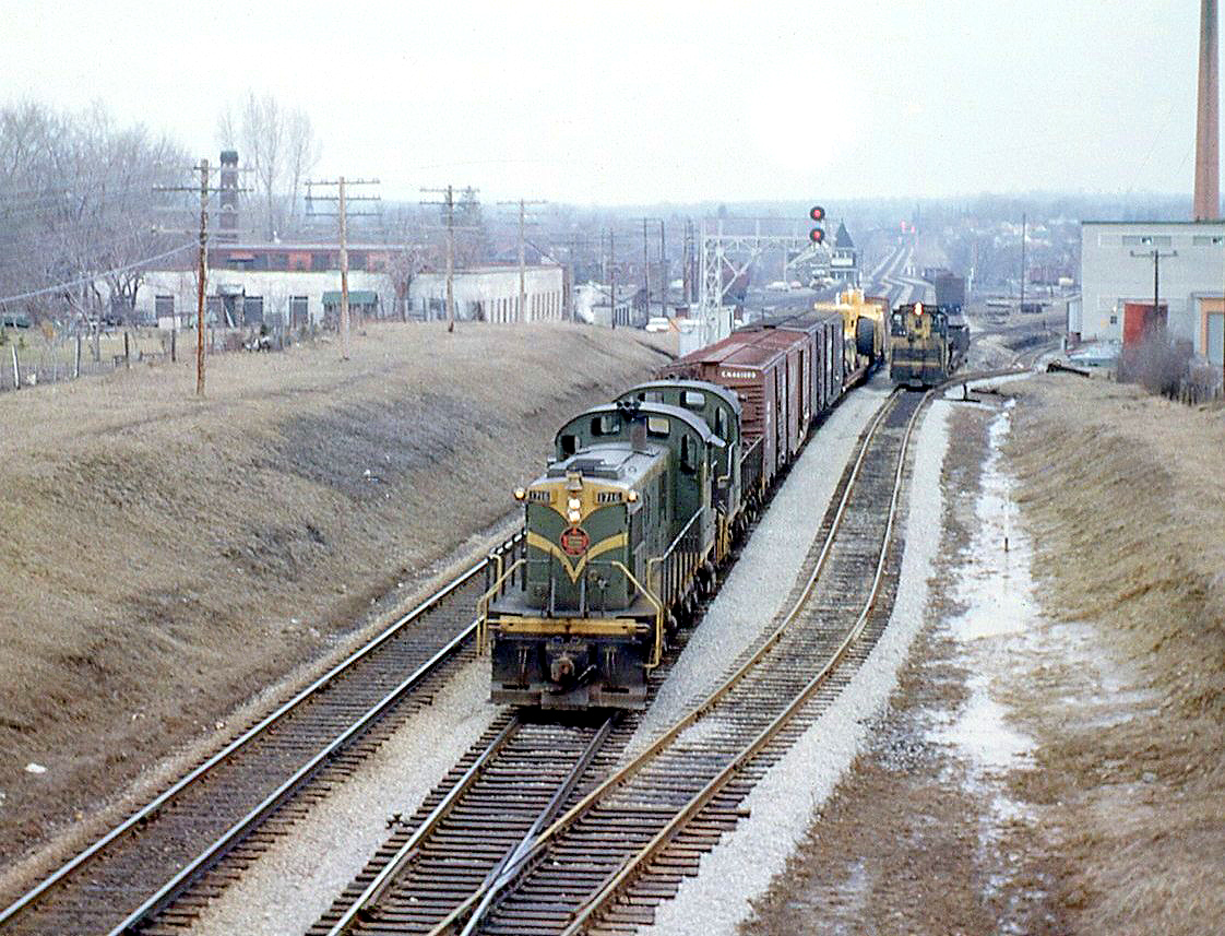 A pair of Canadian National MLW RSC-13's (lead by 1716) head east with a short train at Georgetown ON, on CN's "new" Halton Sub, which had only existed for a few months since the January reorganization. A few cars back is a large dimensional load, which appears to be an earthmover known as a scraper. To the right, a GMD SW1200RS works on the lead to the local Georgetown industries and yard, today used by GO Transit to store and load trains.

The pointed spiral of the station is visible in the background above the train, and further in the distance are the signals for Silver, where the Guelph Sub (formerly part of the Brampton Sub just a few months earlier) branches off from the Halton (made up of parts of the former Brampton and Milton Subs, plus a new section to Toronto (MacMillan) Yard).