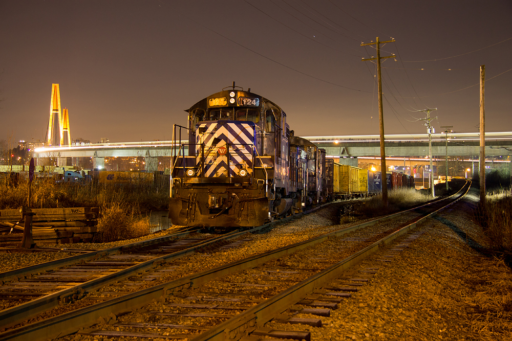 Southern Railways Shops Job sits awaiting a new crew, as a Skytrain streaks by above. Once on duty, the crew will to take the train and head back towards Surrey Fraser Docks where customers like Catalyst await box cars that will be loaded with paper products. At the other end of the train a caboose will be used as a riding platform for one of the crew to spot unprotected crossings.