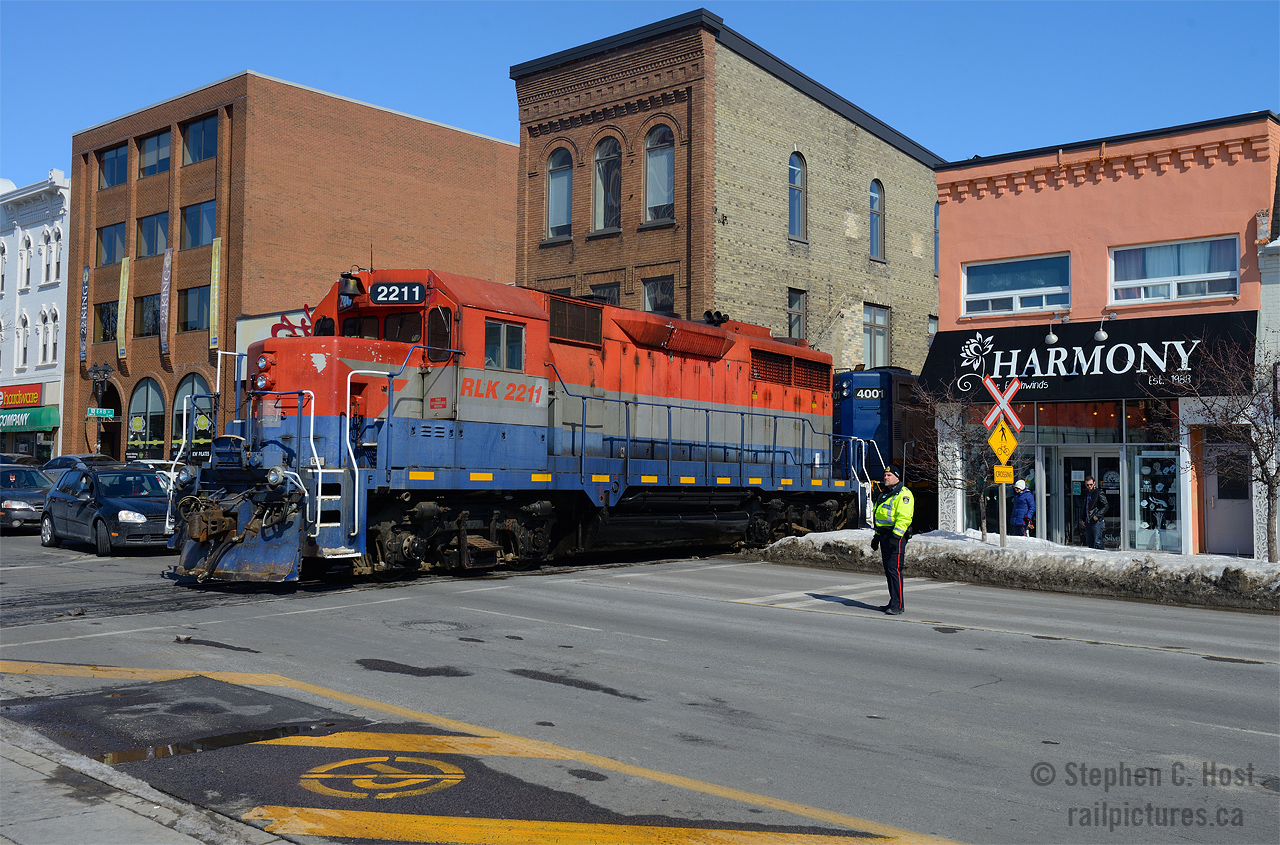 GEXR Train 581-08 making a rare daylight appearance on the Waterloo spur (Kitchener to Elmira). Due to the construction of the Light Rail Transit line, called "ION" - using the Waterloo spur from this point northward, a Welded Rail Train was brought to town. The drop was managed by GrandLinq, the consortium overseeing construction and operation of the line. Dropping rail from Waterloo Station, along U of W to Northfield drive, this sleepy railway line will be transformed into LRT over the next two years. The train dropped rail all day on Saturday, finishing on Sunday by the early afternoon. In this scene, Waterloo Regional police, who were escorting the train across all crossings, is seen flagging traffic as the power ducks between the downtown Waterloo buildings.

Due to LRT construction, freight trains are restricted from 1200-0400 and daylight moves are by far and large very very rare. Once the LRT is open daylight freights will be history. 

More interesting facts: The LRT will use Caroline Street and King St (behind me to the right) for the in-street portion of the LRT, each street will be 'one way' and one track of LRT. A curve will be right where I am standing (approx) to join the Waterloo Spur. The Waterloo spur from this point where the locomotive is and south - it will remain a freight only line. 
A new layover facility complete with loop tracks is presently being built at Northfield Drive in Waterloo to store and maintain the Bombardier Flexity LRT cars (exact same as TTC, just blue). Rails for this yard are part of this drop. The LRT will run all the way to Fairway mall near Cambridge, from there change to BRT to Cambridge. The route is too complicated to type here, maps can be found here - this LRT will transform Waterloo Region and will be a bellweather for other LRT projects in Canada. http://rapidtransit.regionofwaterloo.ca for more.