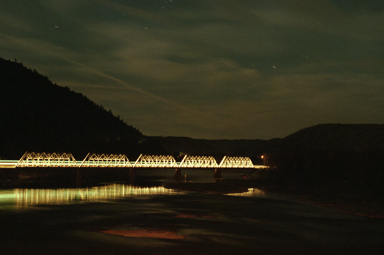 Under a starry sky, the headlight of the westbound Ocean illuminates the rail bridge over the Ristigouche River as it slows for a station stop at Matapédia. The shore to the left is New Brunswick, to the right is Québec.