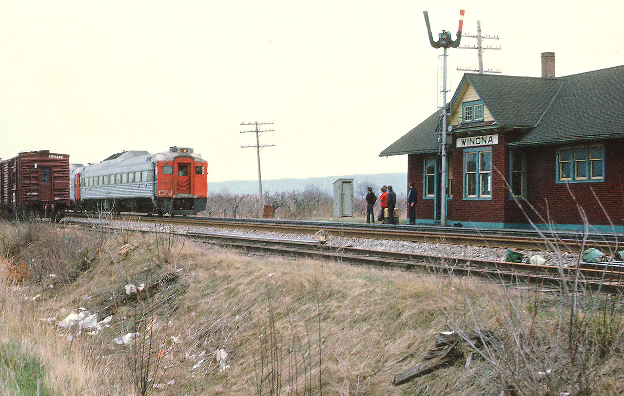 A lot can be said about this image........so...........The trash. Disgusting, but what can you do? Some of us on this group can recall days when trying to photograph stopped trains (or even moving ones!) and just as the shutter clicked, someone on board was throwing garbage out the window. It was almost an epidemic. Metal water cans (10 oz) were the worst. Then it became plastic water bottles. And everything else. This image is an example of that. Wind from the NW has blown collective trash into the ditch.  There are work cars on the North siding by the old Winona station as the CN Budds, Toronto-bound, slow to pick up passengers. One has to wonder if anyone used that classic outhouse in the photo, other than maintenance crews. There were times when I used to see them everywhere along the railways.  They're all gone, and they're certainly not in the hands of collectors of rail artifacts. :o) Probably torched.
Winona station burned down in the fall of 1975. It was located just to the east of Winona Rd; and west of this crossing fruit storage building(s) stood and sidings were usually occupied by cars left in order the locals could fill them with baskets of various crops to rush to the Toronto market. It was once very busy along the line in Niagara, with fruit storage buildings at every community. Naturally, this traffic was eventually lost to trucking, and the daily seasonal local ceased to exist.