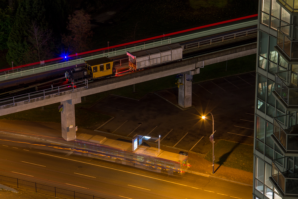 During 2014 Vancouvers Skytrain system saw a power rail upgrade, which caused service to be reduced later in the evenings. One example was this machine, which hauled a tool car and was positioned nicely for some photo opportunities, including a ghostly bus passing through. In the background the lights of a train can be seen streaking on by. This photographer has been fortunate enough over the last 3 and a half years to live right at the end of the line, in a condo overlooking the guideway and King George Station.