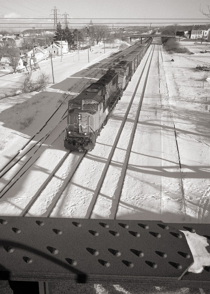 On a sunny January day, we are looking east from the former NS&T bridge over the CN Grimsby sub, as a Hamilton bound freight passes. The train has just passed under the old Merritt Street bridge.  The rail bridge carried the Trillium Rail Town Line Spur, now out of service, the span has been removed.
