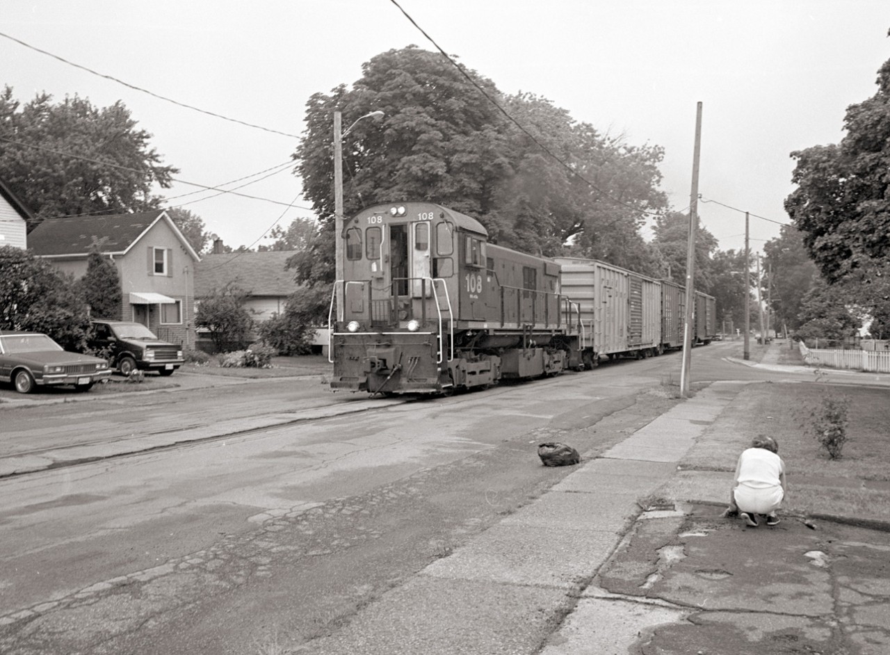 Trillium Railway 108 makes its way up the Townline Spur on its way to Interlake Paper, past a lady tidying her garden who doesn't give the train a second look.  The locomotive, a former CN S-13, built by the Montreal Locomotive works in 1959, is still active but the street running is no more.