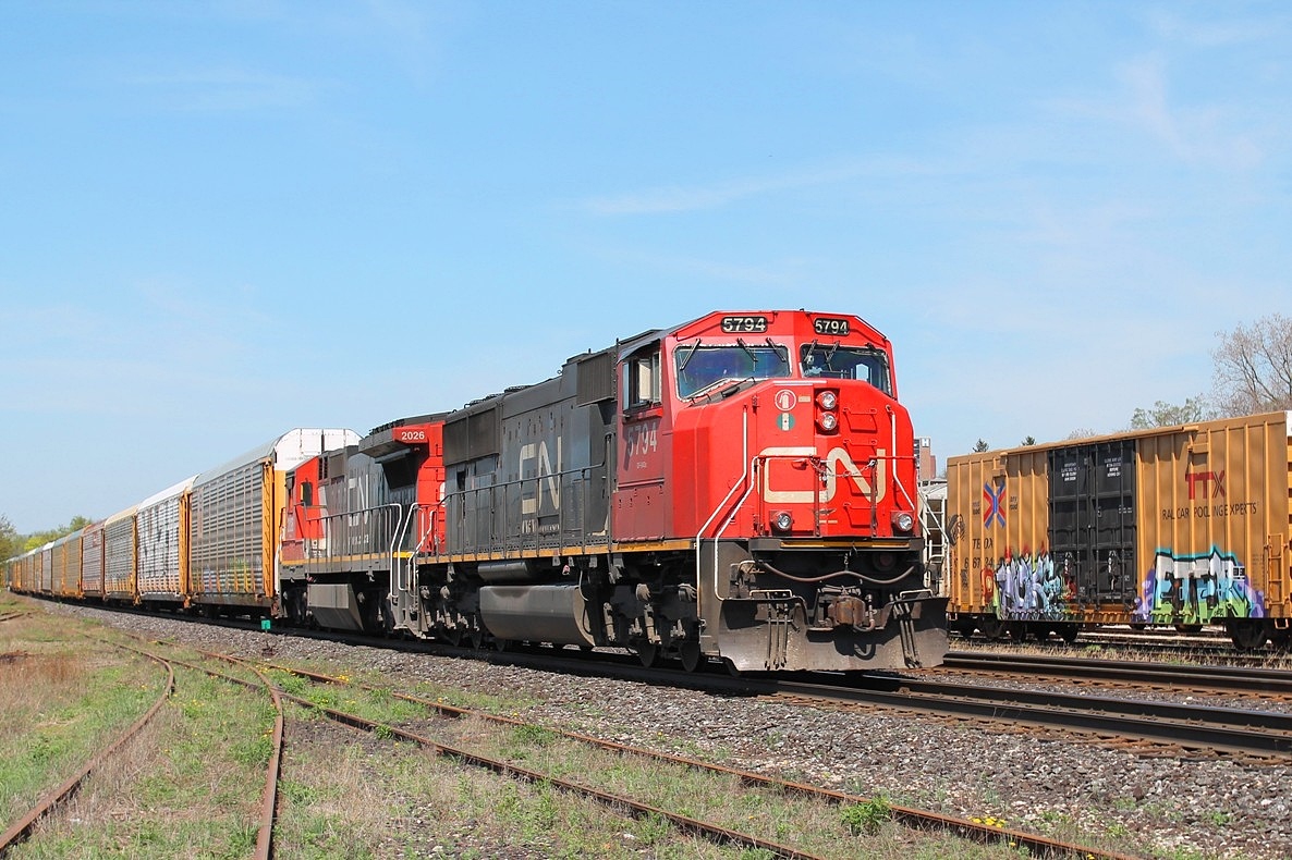 On the hottest day of the year so far, CN 5794 and 2026 lead an eastbound through the yard. They are passing a line of box cars that have suffered from illicit art!