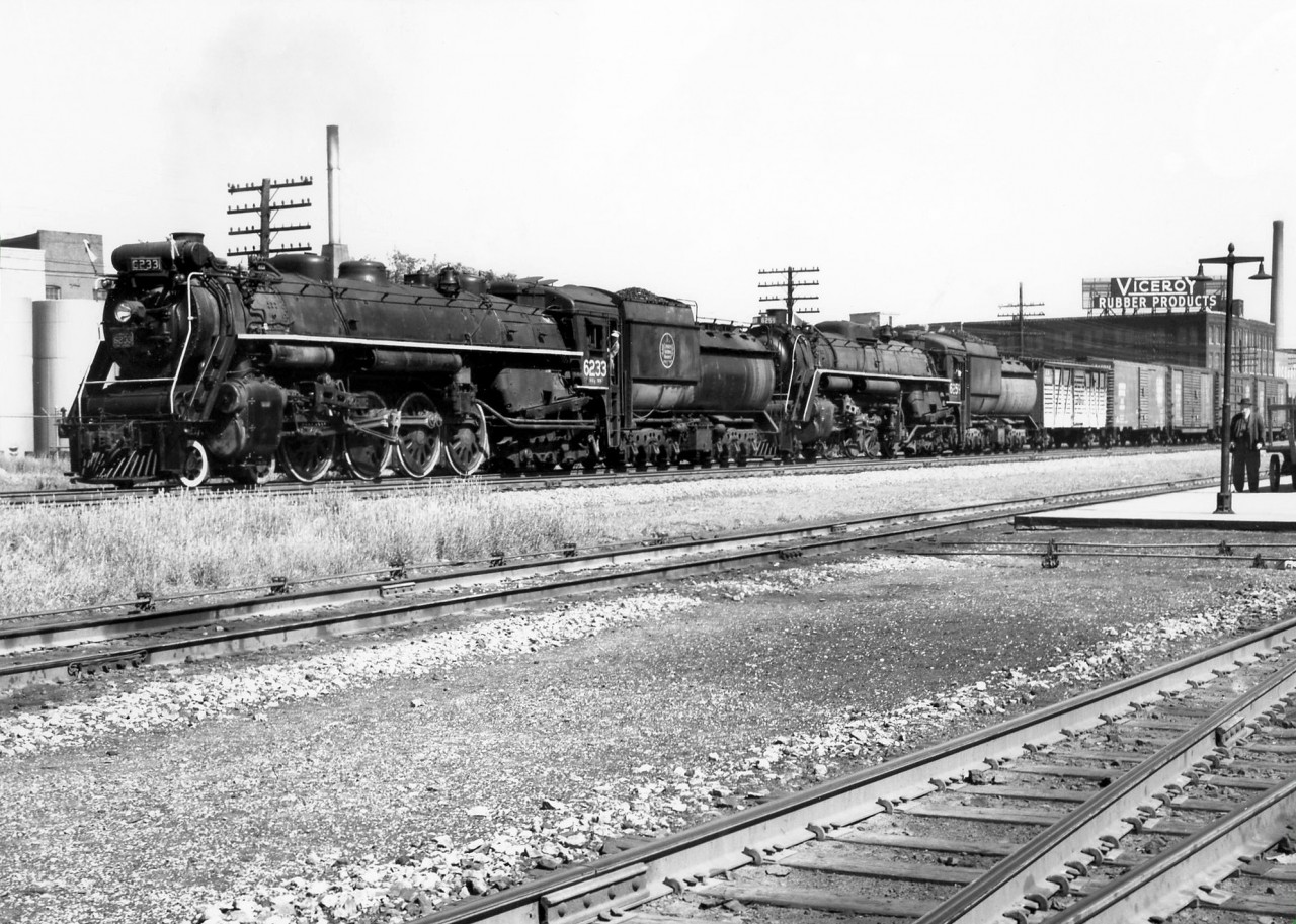 Doubleheading CN Northerns 6233 and 6259 wait to be cleared to cross the diamond northbound on the CN Brampton Sub (later renamed the Weston Sub), at West Toronto Junction on a sunny June day in 1958 (about 16:00 hours). In tow is a freight with a single stock car visible behind the power, and single and double door boxcars, the latter of which was still the primary means of transporting automobiles by rail. On the right an individual and baggage cart on the platforms of CPR's West Toronto Station can be seen, and the Galt Sub. In the background is the old Viceroy Rubber factory off Dupont Street.  At the time, the linkage rods along the Galt Sub track and by the station that control signals and switches signify the diamonds were still controlled by the old West Toronto interlocking tower just to the north, visible *here*. A few years later in 1965, the whole area would be converted to CTC, controlled by CP, and the tower demolished. A view of the diamonds then can be seen *here*. Today, GO Transit MP40's hauling bilevels, VIA corridor trains and Union Pearson Express DMU's replace the CNR Northerns, freight trains, GMD1-hauled commuter trains and RDC's of yesteryear on the Weston Sub.