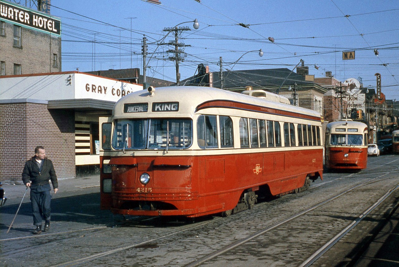 A pair of Toronto Transit Commission PCC cars wait at the start of The Queensway to turn into the TTC's Roncesvalles Carhouse, at the intersecting junction where King Street, Queen, Rocesvalles and The Queensway all come together in an interesting arrangement. Car 4215 was out on a King run, and 4232 behind signed up Queen. Another PCC is also visible along Queen St., lined with storefronts advertising for CIBC, the Royal Bank of Canada, and Pickin' Chicken BBQ.

Behind the 4215 is Gray Coach Lines' Sunnyside bus terminal. GCL was an interurban coach operator owned by the TTC, with many routes branching out from Toronto to places as far as London, Owen Sound, and North Bay, advertising "Travel The King's Highway" and other travel information in neon lighting on its facade. Part of the nearby Edgewater Hotel's signage (today a Howard Johnson) is visible on the left behind the GCL terminal (now a McDonalds). The old CNR Sunnyside Station is out of view to the right.