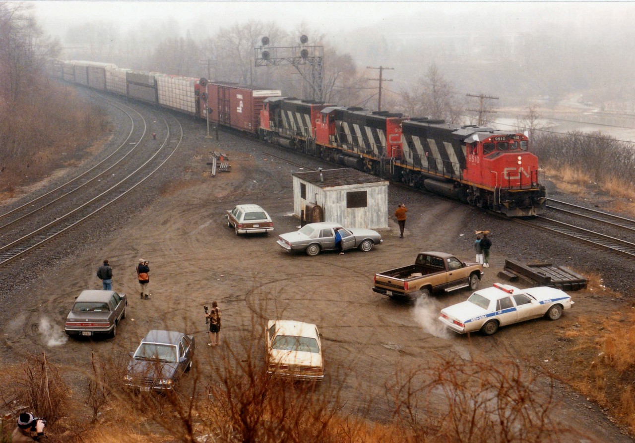 In my opinion, this oddball shot overlooking Bayview Junction is a classic example of what most weekend railfanning was all about. Note it is cold (engines running) and it is miserable (warm coats) and it is damp fog, the worse kind of spring day; but several fans still congregate at the favourite train-watching spot. The Hamilton police officer just drops by from time to time to shoot the breeze and pick up tidbits of information he might find use for. Very cordial. Although this image is only 26 yrs old at this time of posting, it shows how the guys would get together in order to exchange information. No computers, no cell phones. Often most of the day would be spent here, as the CN 9510, 9513 & 9503 is Niagara-bound and the track to the left is the main line out to London/Sarnia, etc. This was the place to be for action, even though it didn't take long to run thru all the "photo angles" there might be.  Of course this is all changed now, fenced, off-limits, and good times here but a memory. Fans who still show at Bayview use the walkbridge over the tracks, just barely visible in the fog, extreme upper left. My car? The station wagon up by the shack. The usual blue heap was not on the road that day, having died for about the 50th time. It needed crutches, not tires.