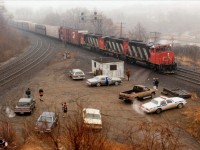 In my opinion, this oddball shot overlooking Bayview Junction is a classic example of what most weekend railfanning was all about. Note it is cold (engines running) and it is miserable (warm coats) and it is damp fog, the worse kind of spring day; but several fans still congregate at the favourite train-watching spot. The Hamilton police officer just drops by from time to time to shoot the breeze and pick up tidbits of information he might find use for. Very cordial. Although this image is only 26 yrs old at this time of posting, it shows how the guys would get together in order to exchange information. No computers, no cell phones. Often most of the day would be spent here, as the CN 9510, 9513 & 9503 is Niagara-bound and the track to the left is the main line out to London/Sarnia, etc. This was the place to be for action, even though it didn't take long to run thru all the "photo angles" there might be.  Of course this is all changed now, fenced, off-limits, and good times here but a memory. Fans who still show at Bayview use the walkbridge over the tracks, just barely visible in the fog, extreme upper left. My car? The station wagon up by the shack. The usual blue heap was not on the road that day, having died for about the 50th time. It needed crutches, not tires.