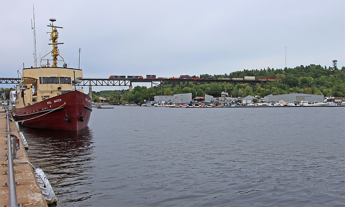 A CN stack train heads over Parry Sound harbor with MV Still Watch still watching. The ship was built in 1960 as a research vessel for the Canadian Coast Guard and is now privately owned.