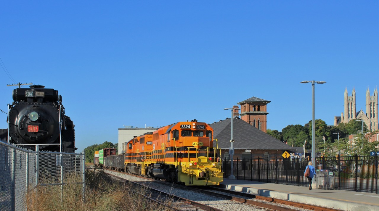 Goderich Exeter 3394 leads Goderich Exeter 3393 past the Guelph train station in downtown Guelph, as CN 6167 (4-8-4) steam locomotive, sits behind its fenced in resting spot watching on. The cities main feature, the Church of our Lady, is at the right.