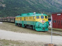 The three foot gauge White Pass and Yukon Railway was originally built to provide transportion toward Yukon's Klondike Gold Rush area from the nearest port, Skagway Alaska. Nowadays, it mainly serves as a summer-season passenger railway carrying cruise ship and other tourists to view the mountain scenery on a round trip from Skagway Alaska to the White Pass Summit, or the longer trip between Skagway and Fraser BC (about 12.5 km into Canada, where the Klondike Highway comes close to the railway).  <br> <br>
GE shovelnose units WPY 91 and 97 are running around their train at Fraser BC, for the return trip to Skagway Alaska.  Passengers from a tour bus can be seen going to board the train.  The Fraser station sign indicates 27.7 rail miles to Skagway.  Note the portable railway crossing sign, weighed down by a small flanged wheel and rail joiners. <br> <br>
Originally powered by Alco 6-251 prime movers, these locomotives have been re-engined with Cummins QSK45L diesels and now sound totally different, much higher revving.  I miss the Alco chug, but lower emissions is an important benefit.