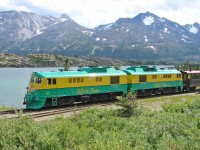 Nowadays, three foot gauge White Pass and Yukon Railway mainly serves as a summer-season passenger railway  carrying cruise ship and other tourists to view the mountain scenery on a round trip from Skagway Alaska to the White Pass Summit, or the longer trip between Skagway and Fraser BC (about 12.5 km into Canada, where the Klondike Highway comes close to the railway). <br> <br>
GE shovelnose units WPY 97 and 91 are northbound with their train past the south siding switch at Fraser BC, with Bernard Lake alongside. Passengers arriving by tour bus will board at Fraser, for their return to Skagway. <br> <br>
Originally powered by Alco 6-251 prime movers, these units have been re-engined with Cummins QSK45L diesels and now sound totally different, much higher revving.  I miss the Alco chug, but lower emissions is an important benefit.