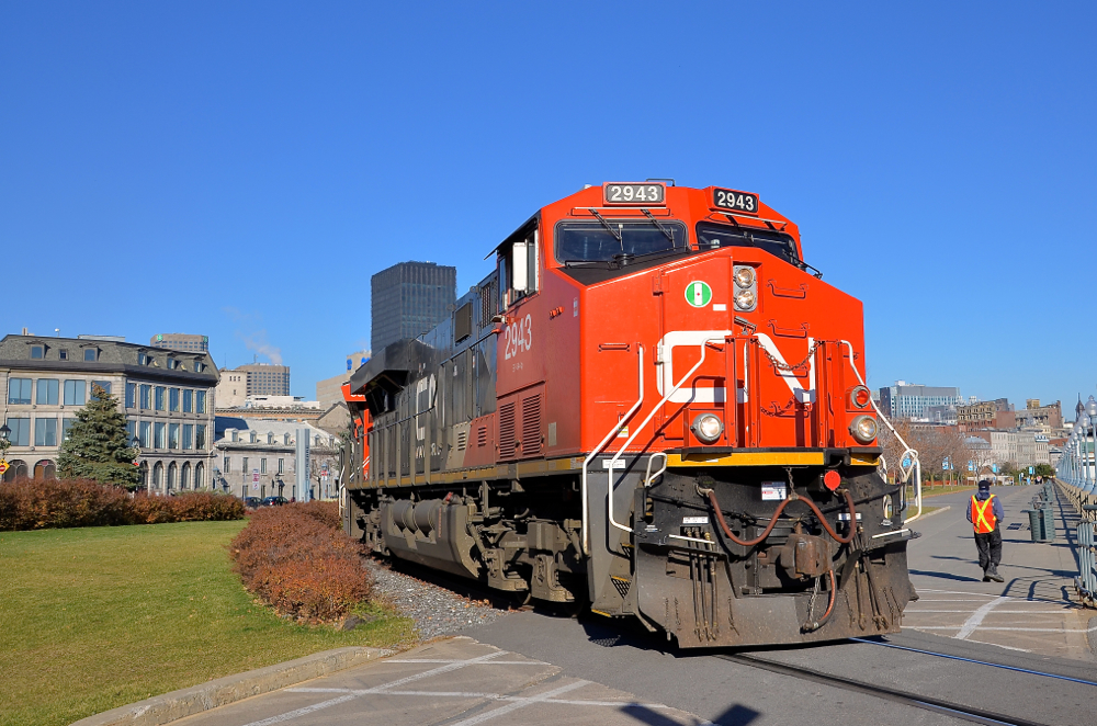 Flagging the crossing. CN 2943 is in charge of CN 149 as it rounds a curve in the Port of Montreal, with CN 3023 trailing. In a few seconds the head end will cross the Lachine canal on a sunny fall morning. At right is a Port of Montreal employee flagging this crossing.