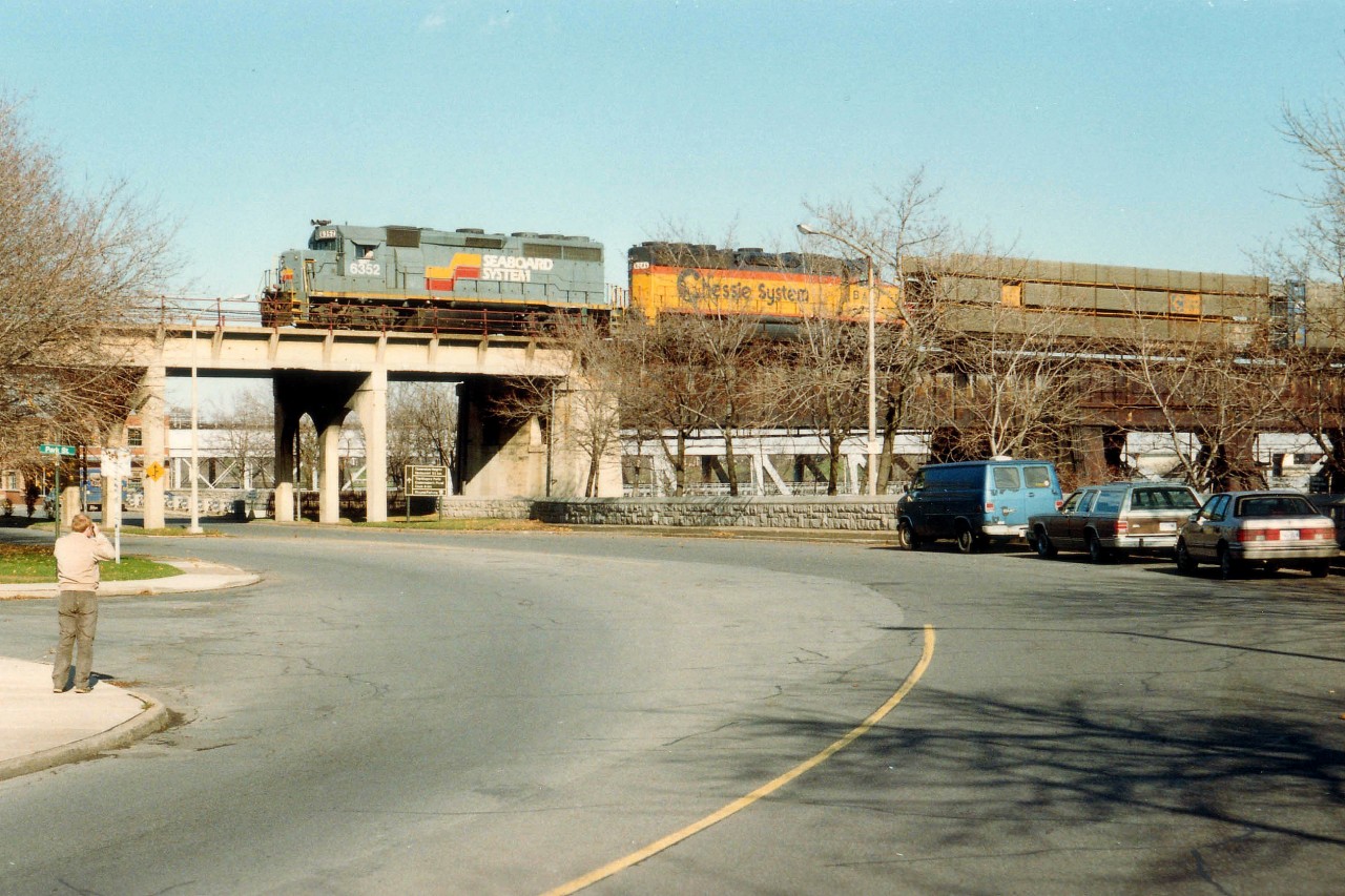 An early afternoon CSX tri-weekly #321 rolls into Canada by way of the now abandoned Steel Arch Bridge spanning the Niagara River. Power is CSX 6352 and 6246; the leader looking spiffy in that old Seaboard paint scheme. It has been a few years, but I believe the photographer on the sidewalk is one Dave Howard, as he appeared before the wife eventually fattened him up. :o) For members of this list who might be curious, as in "Snake", the middle car (Colony Park Wagon) on the right is what I was driving. The Blue machine was down for repairs.