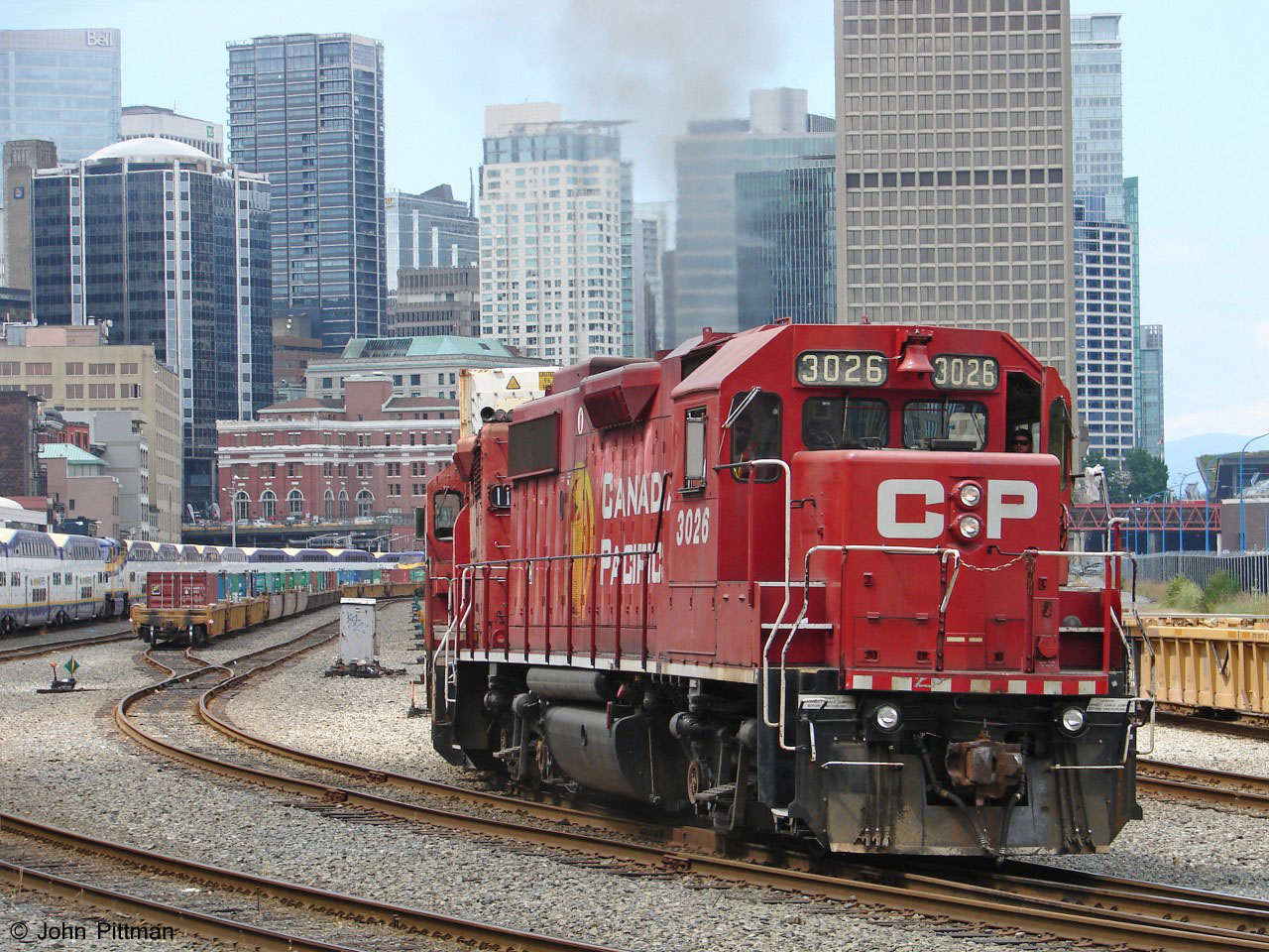 GP38-2 CP 3026 drags control cab CP 1126 (ex-GP35) and a long string of well cars east out of a siding in the CP station yard in downtown Vancouver. Track ends within a mile west, under the Waterfront Centre.  
The red brick building is the West Coast Express Waterfront commuter station, formerly CP Vancouver station. W.C.E. trains can be seen tied up on the left side, awaiting their evening commuter runs.