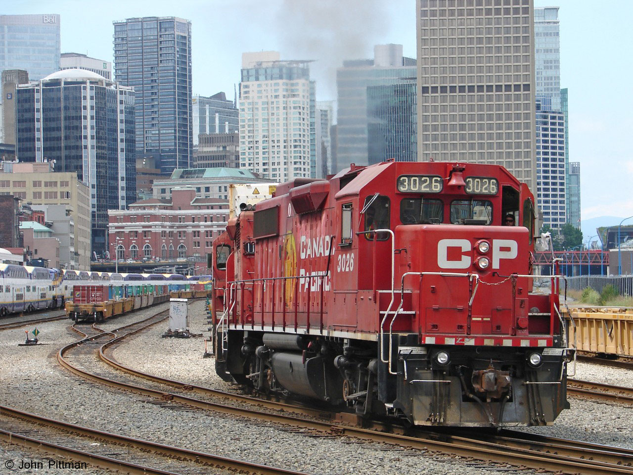 GP38-2 CP 3026 drags control cab CP 1126 (ex-GP35) and a long string of well cars east out of a siding in the CP station yard in downtown Vancouver. Track ends within a mile west, under the Waterfront Centre.  
The red brick building is the West Coast Express Waterfront commuter station, formerly CP Vancouver station. W.C.E. trains can be seen tied up on the left side, awaiting their evening commuter runs.