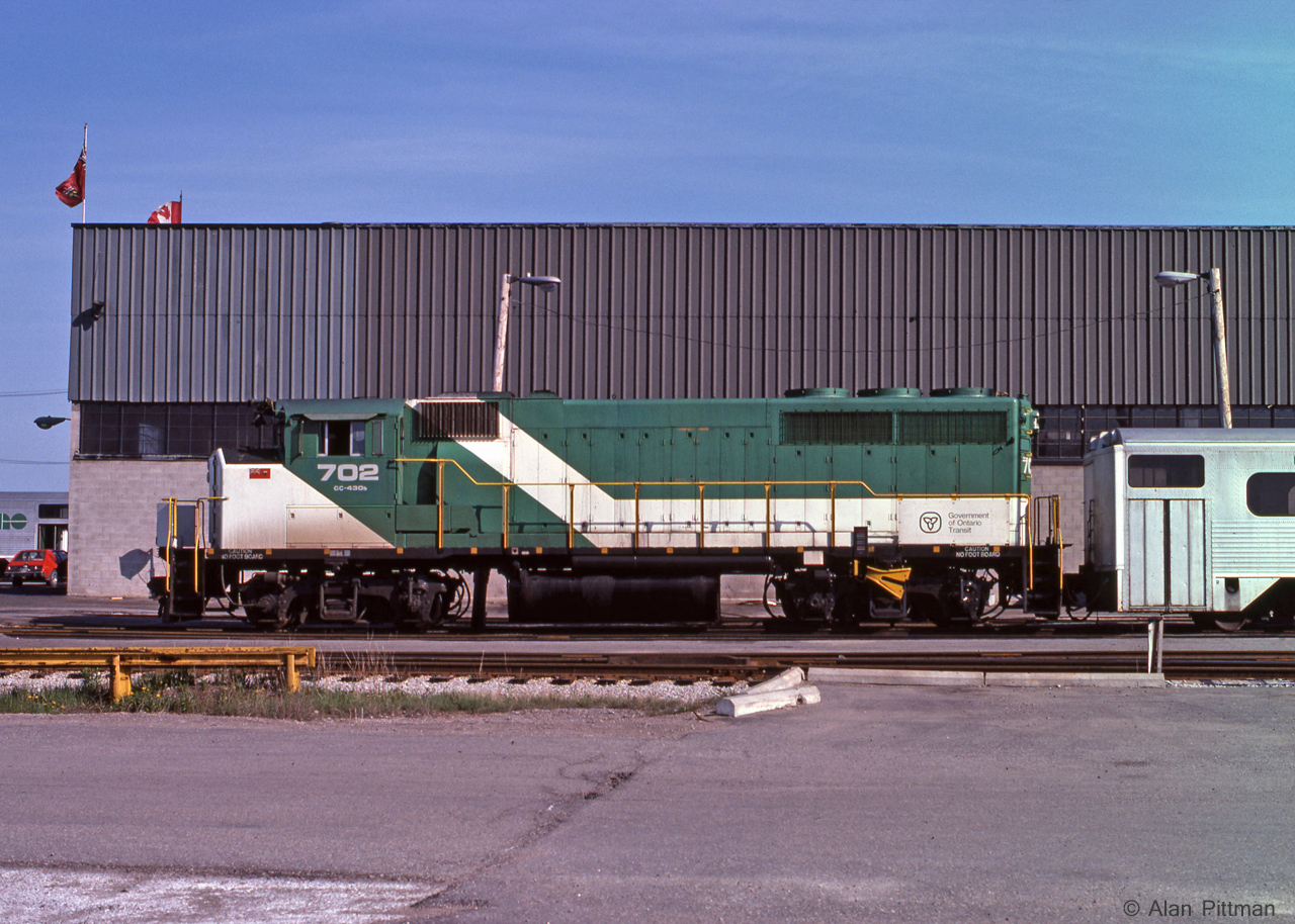 GP40-2(W)  GOT 702 is tied up outside GO Transit's Willowbrook maintence facility this June 1978 weekend, with a train of GO Transit's original single-level coaches.  At the front of the building can be seen another coach and an AMC Gremlin.
This loco was delivered to GO Transit in 1973 as GOT 9810, and was re-numbered GOT 702 in 1975. Unlike GO's previous locomotives (GP40TC), units 700-710 had no Head End Power (HEP) generation for passenger coaches. They were intend to be used with an APCU (GO 900-911) unit at the other end of the train. (Other HEP alternatives being an ex B-unit APU's, or with an HEP equipped locomotive).
Subsequently GO mainly bought HEP equipped locomotives, and the GP40-2(W)'s were sold in 1991, all but one going to CN.  GO 702 became CN 9670; the ex-GO units ran in Green and White paint on CN for a while.
