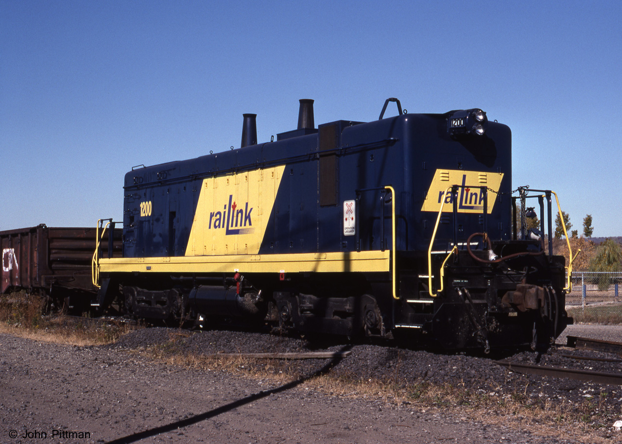 Only one of its kind in Canada, RaiLink painted B-unit switcher 1200 was having a rest while a pair of ex-CN SW1200RS were switching cars in Hamilton's Stuart Street yard. 
Originally built as IC 9203B in Feb 1940, it was the cabless B-unit half of an EMD "TR" switcher pair - often nicknamed "cow and calf". Both parts of the TR were based on the EMD NW2, and it originally had the NW2-style dip in the hood in front of the (absent) cab. IC renumbered it to IC 1026B in 1956. 
In 1998 this locomotive was 58 years old - sort of. I expect the frame and some other parts were that old. But IC had rebuilt it into an "SW13B" in 1972, with many parts replaced, the hood line flattened, and re-numbered to IC 1300B.  Later becoming ICG 1300B, it passed to the Paducah & Lousville as PAL 1300B. 
The unit was acquired freshly painted for RaiLink Hamilton in 1998, and as I recall was active for at least a couple of years. My 2005 Cdn Trackside Guide lists it in [brackets] indicating stored unservicable. Mark Macauley posted an image of it from October 8 2008 when scrapping was in progress  - http://www.railpictures.ca/?attachment_id=21772