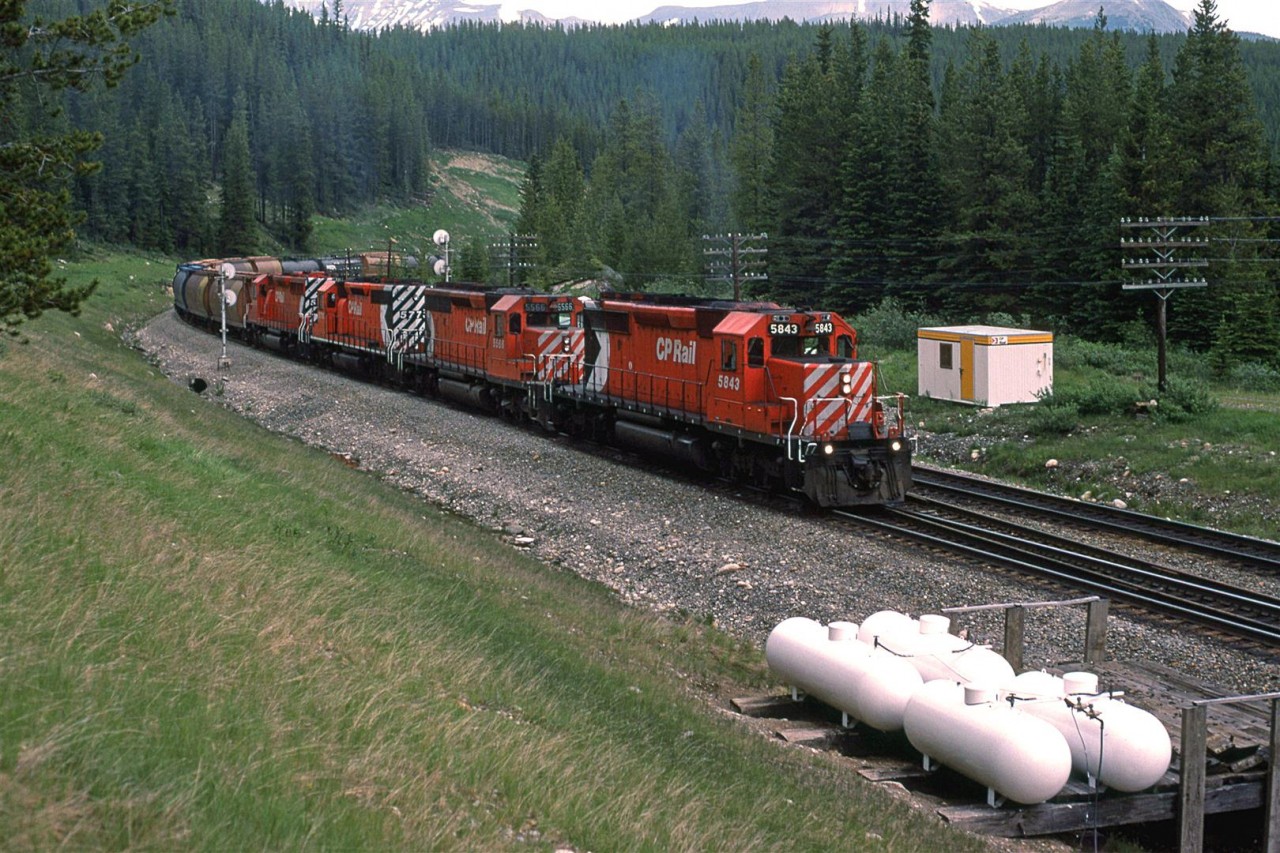 This westbound grain train is very close to the high point of Kicking Horse Pass. The propane heaters are for the crossovers for a grade separation between this point and Lake Louise. The train is on the new gentler alignment, while the old line is obscured by the train.