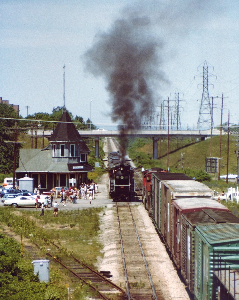 Its a rather eventful Saturday for the fans gathered at the old Grimsby CN station as CN 6060 starts up eastbound as a westward CN freight crawls by. This special oil-fired (converted) locomotive never failed to attract a crowd. That is the Christie St overpass in the background, and I am standing, alone, I might add, on the Maple Av bridge, with most fans content with being at the station. The track in the foreground veers off to the historic GTR station (out of view). The track looks disused in this scene; last time I saw anything on it was a boxcar a few months earlier, probably a bad order car. The track was removed a few years after this image was taken, and the CN station burned down in an electrical fire on the very last day of 1994. Currently a VIA kiosk occumpies this location, yet only Amtrak provides service down thru Niagara any more.