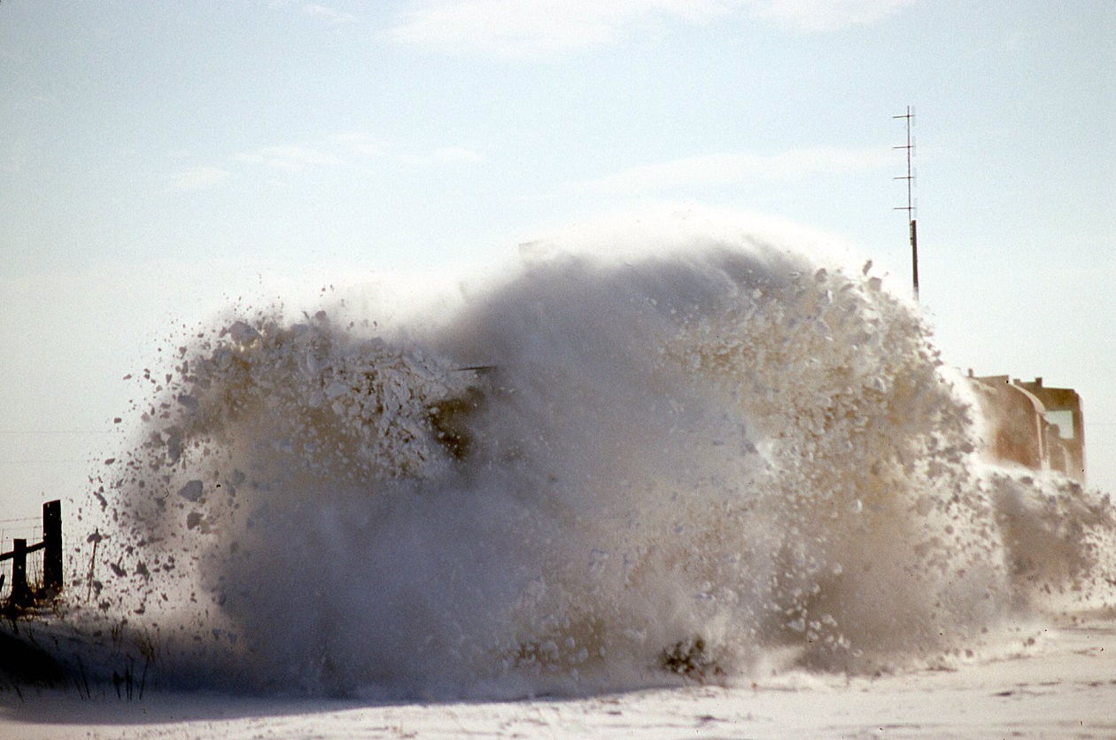 A frozen drift of heavily packed snow explodes as the plow extra north hits it hard at speed.