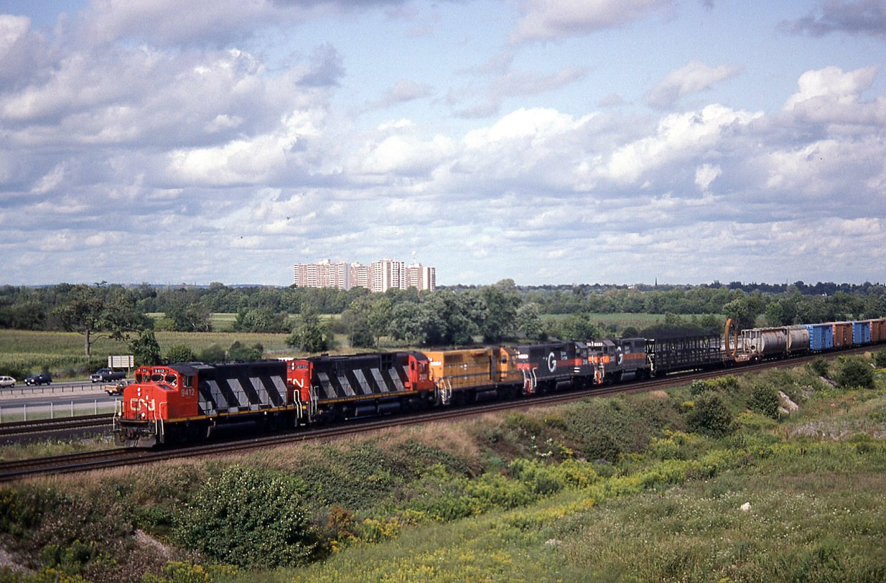 CN 9472,2031 with MEC 256, B&M 258, 263 dead in tow wb just about to duck under Durham 23 the Ajax, Whitby boarder. Destination of the last three units is unknown. Note the first two cars next to the units. A snow fence multi and a depressed flat carrying a big metal zero of some type. This location is now a total mess at this time. A new bridge for Durham 23 and a new interchange for the 412 ? to connect to the 407 extension.