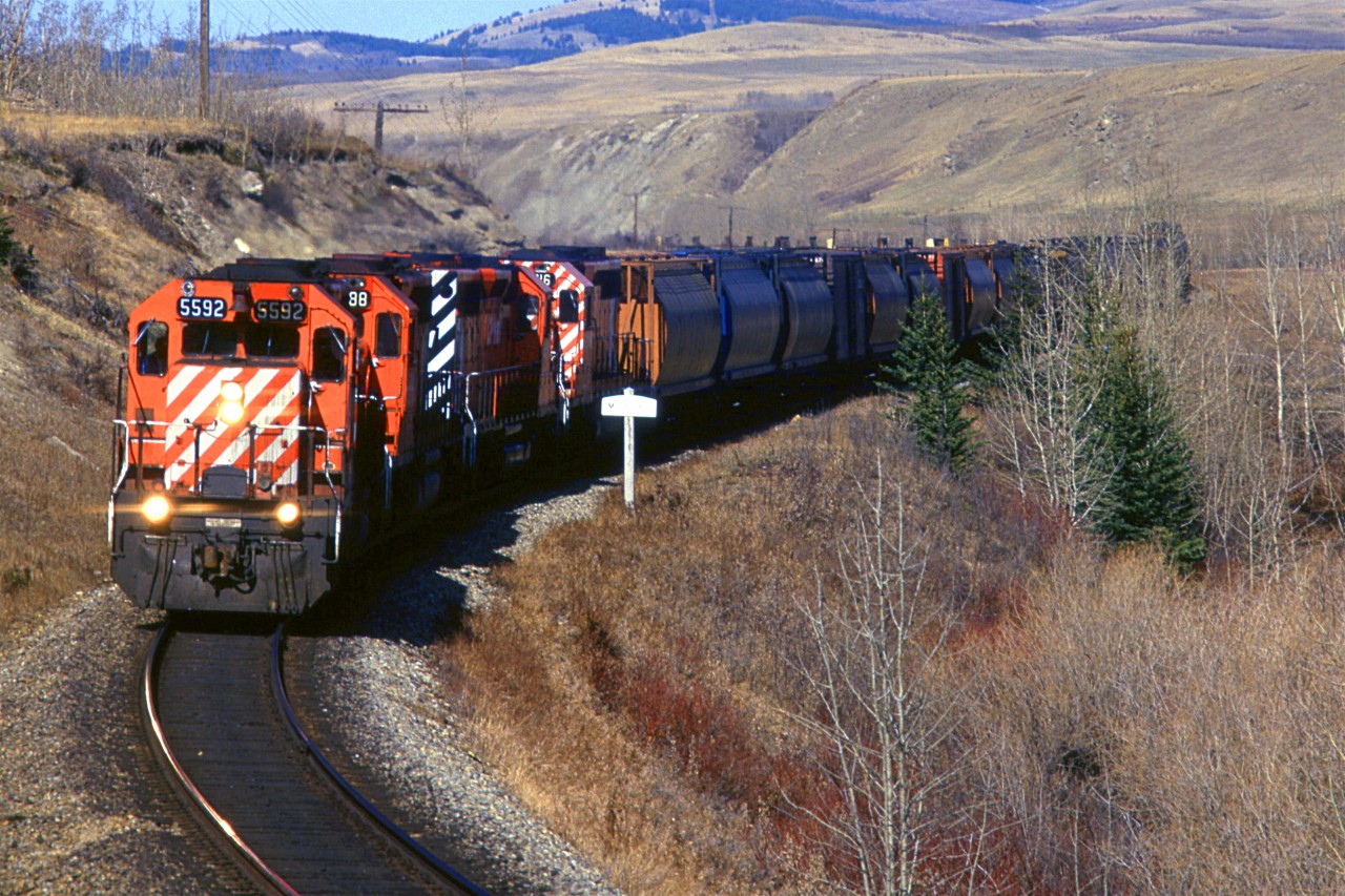 A westbound empty grain train is by the sign indicating that the next siding/station is Mitford. This is west of Cochrane in the Bow River valley.