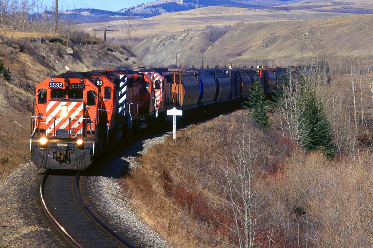 A westbound empty grain train is by the sign indicating that the next siding/station is Mitford. This is west of Cochrane in the Bow River valley.