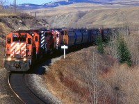 A westbound empty grain train is by the sign indicating that the next siding/station is Mitford. This is west of Cochrane in the Bow River valley.