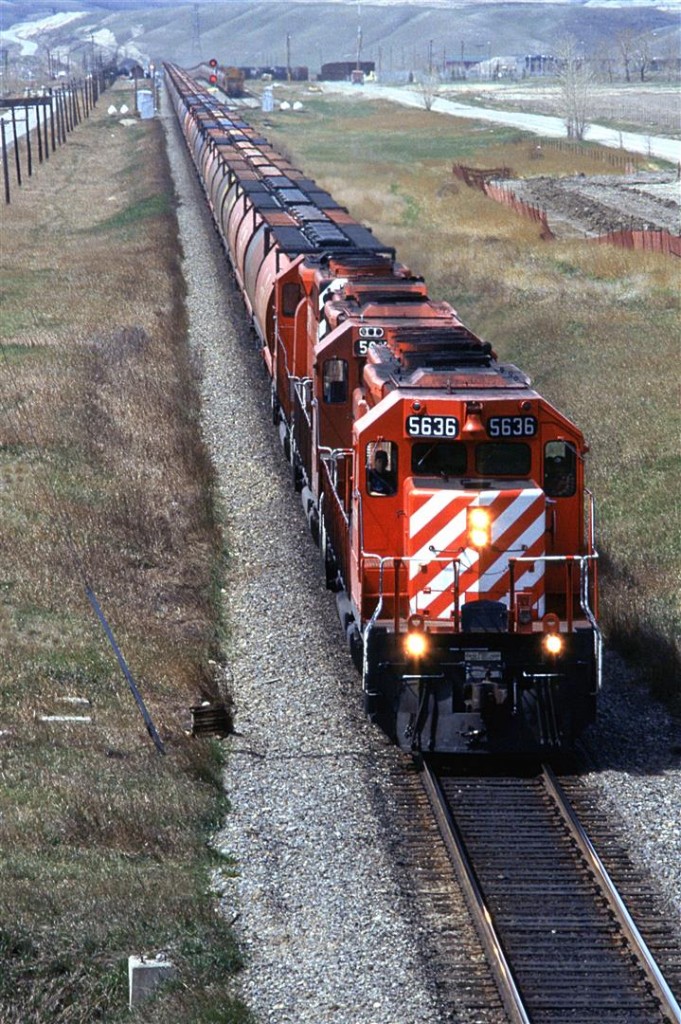 An eastbound empty grain train passes through Kieth yard, in the background. What looks like snow in the background is gravel. There is a large aggregate operation on/in that hill on the horizon.